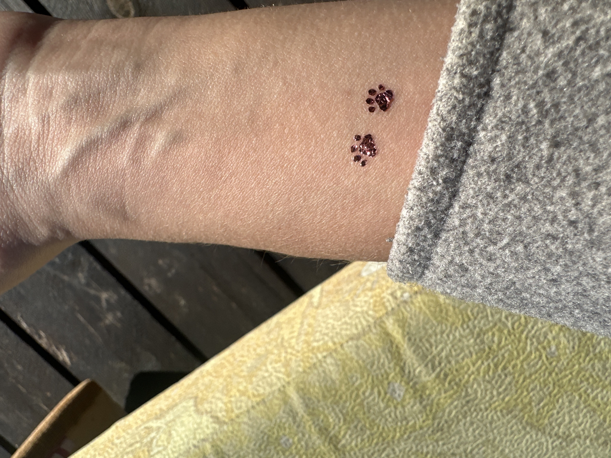 The image shows a person's forearm with two glittery paw print designs on the skin. The forearm rests on a surface covered with a yellow, textured fabric, and a portion of a gray sweater sleeve is visible. A dark, wooden surface with planks is visible in the background. The location is not discernible from the image and cannot be identified as Kharnavy, Russia.