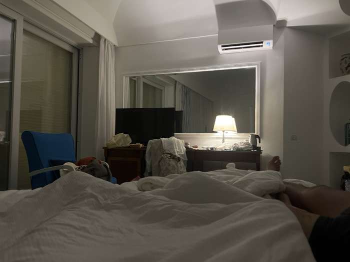 This image presents a detailed view of a bedroom interior, located in Massa Lubrense, Italy, captured from a low vantage point, likely from a bed. The foreground is filled with white, textured bedding. On the right side of the foreground, the bare legs and feet of an individual are visible, resting on the bed.
To the left, a blue upholstered armchair is positioned near a large sliding glass door or window, which is covered by internal blinds or a reflective surface, and partially obscured by a sheer white curtain. In the mid-ground, a dark flat-screen television sits on a wooden dresser or bedside table. An illuminated table lamp with a white shade stands to the right of the television, casting light and reflecting prominently in a large, rectangular mirror mounted on the wall behind the dresser. Various items, including a black electric kettle, are present on the dresser's surface.
Above the mirror, a white air conditioning unit with a visible blue digital display is mounted on the ceiling. The right wall features several white, built-in arched display niches, one containing a decorative white and blue vase. The room is characterized by uniformly white walls and ceiling, illuminated by artificial light sources, creating a subdued indoor environment.