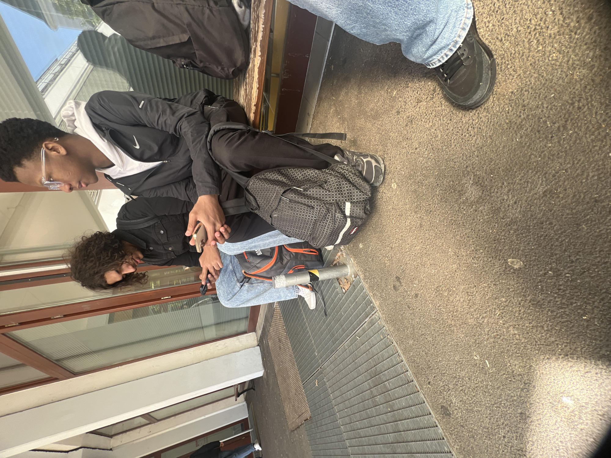 Two individuals are seated on a metal pipe near a building with windows in Schiltigheim, France. The person on top wears a black jacket with a white hoodie underneath, black pants, and carries a black and white patterned backpack. They are wearing glasses and looking down. The other person is wearing a black jacket, blue jeans, and white sneakers. Both individuals are holding cell phones in their hands. A third person is partially visible above them, wearing blue jeans and black shoes. The setting appears to be an outdoor area with a concrete floor and a metal grate. A backpack and a dark-colored bag are present near the top of the image.