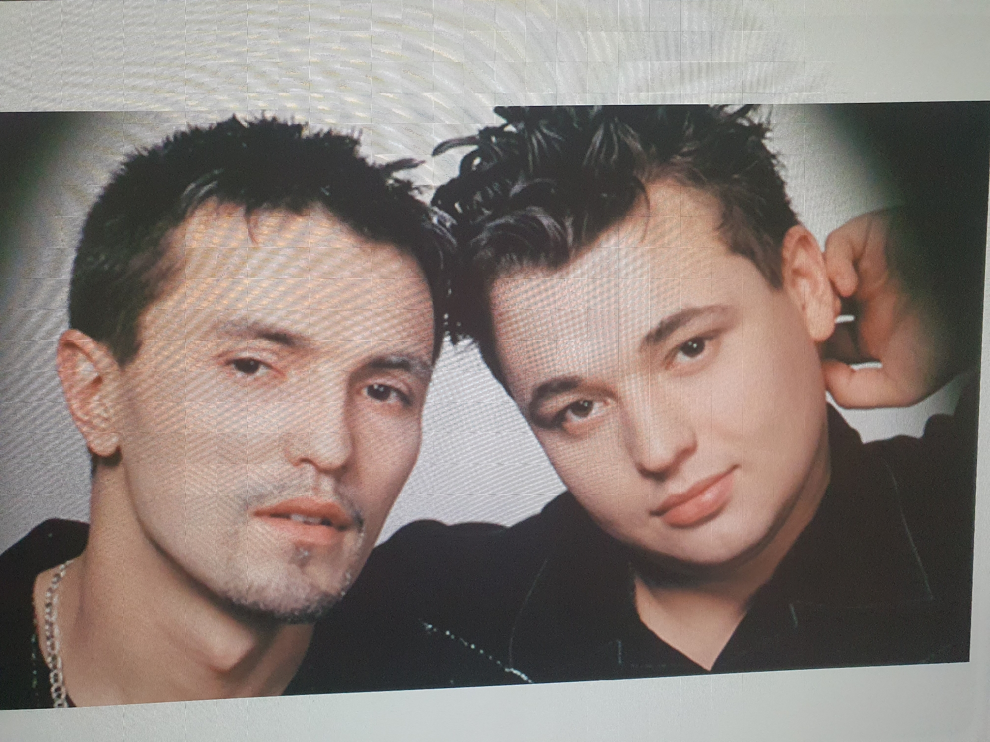 The image shows a close-up, head-and-shoulders portrait of two young men. The man on the left has short, dark, spiky hair, dark eyes, and a light beard. He is wearing a dark shirt and a silver chain necklace. The man on the right also has short, dark, spiky hair and dark eyes. He is wearing a black shirt and has his right hand near his ear. They appear to be posing for a photo. The background is a plain, light gray. There is no indication of location in the city of Tashkent, Uzbekistan.