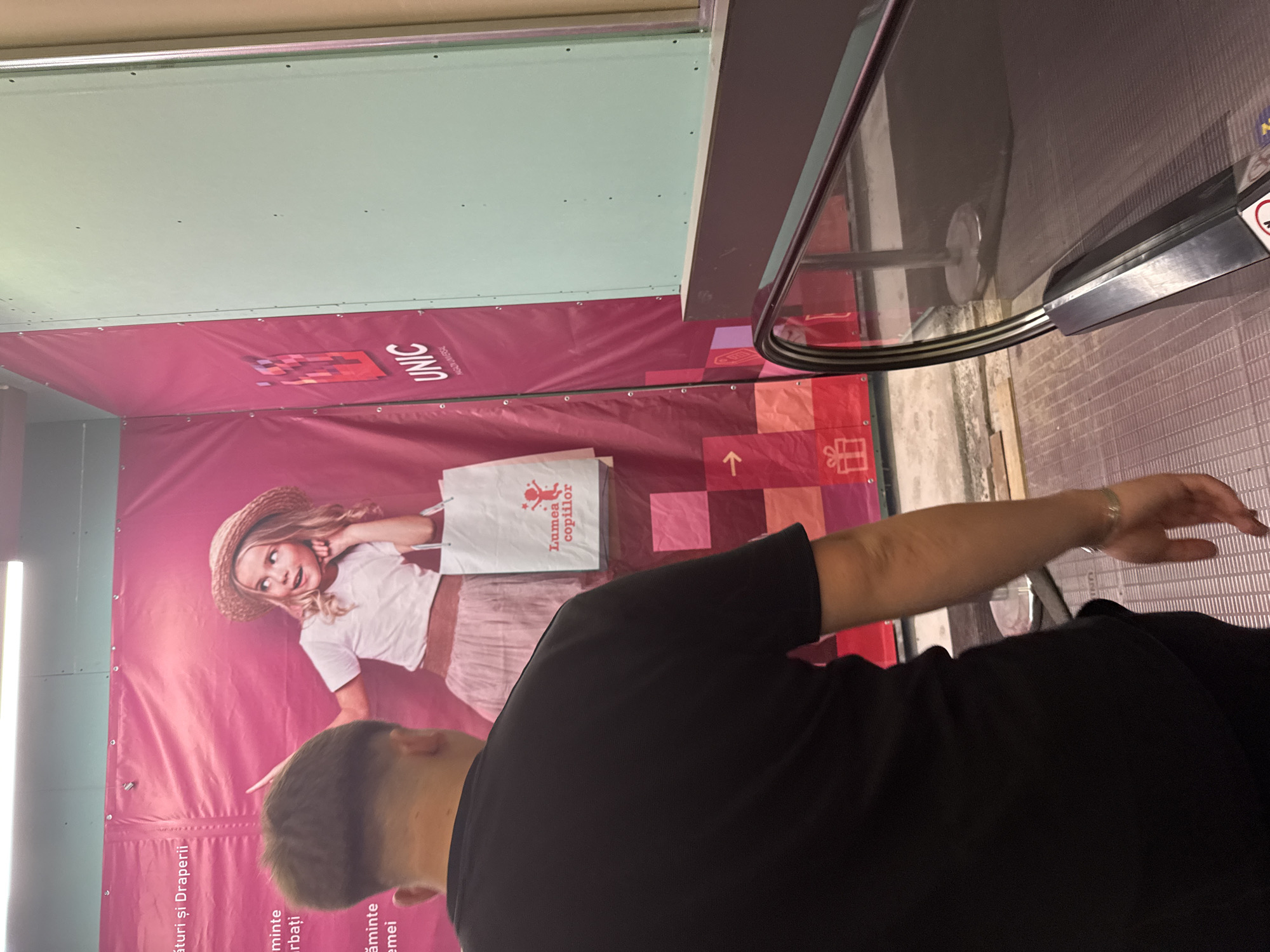 The image depicts a person in a black t-shirt standing on an escalator, with their back to the camera. They are looking towards a large pink advertisement banner. The banner features a young girl in a straw hat and a white shirt, holding a shopping bag with Romanian text that reads "Lumea copiilor" (Children's World). The banner also has the logo and name "UNIC MAGASIN UNIVERSAL" (UNIC DEPARTMENT STORE). This suggests the image was taken at the UNIC department store in Chișinău, Moldova. The escalator is made of metal and has a reflective surface. The environment appears to be indoors, possibly a shopping mall or a department store. There are some construction materials visible on the floor near the banner.