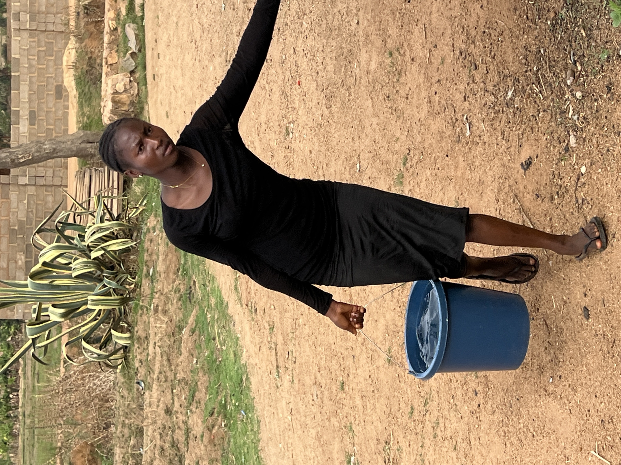 A woman wearing a long-sleeved black dress and sandals is holding a blue bucket filled with water. The location appears to be Kirana, Nigeria, based on the landscape and architectural styles visible. The woman is standing on a dirt path with sparse grass. Behind her, there is a building constructed from concrete blocks, and a plant with yellow and green striped leaves. The scene is outdoors during daylight.