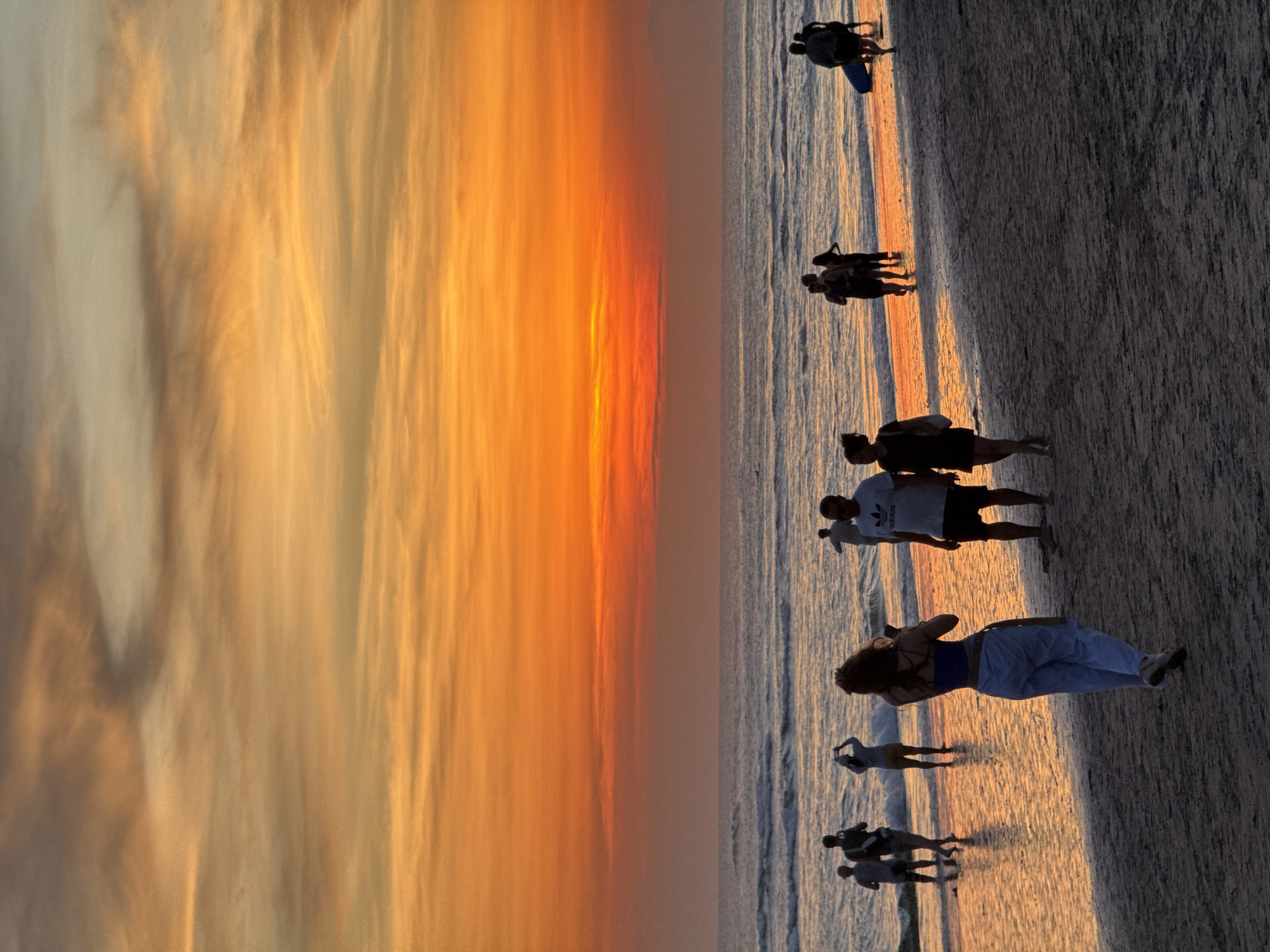 The image captures a sunset scene on a beach in North Kuta, Indonesia. Several groups of people are present, some walking along the shoreline, others gathered in small groups. The sky displays a vibrant gradient of orange and yellow hues, typical of a tropical sunset. The water reflects the colors of the sky. The beach is sandy, and the light from the setting sun casts long shadows of the people onto the sand.