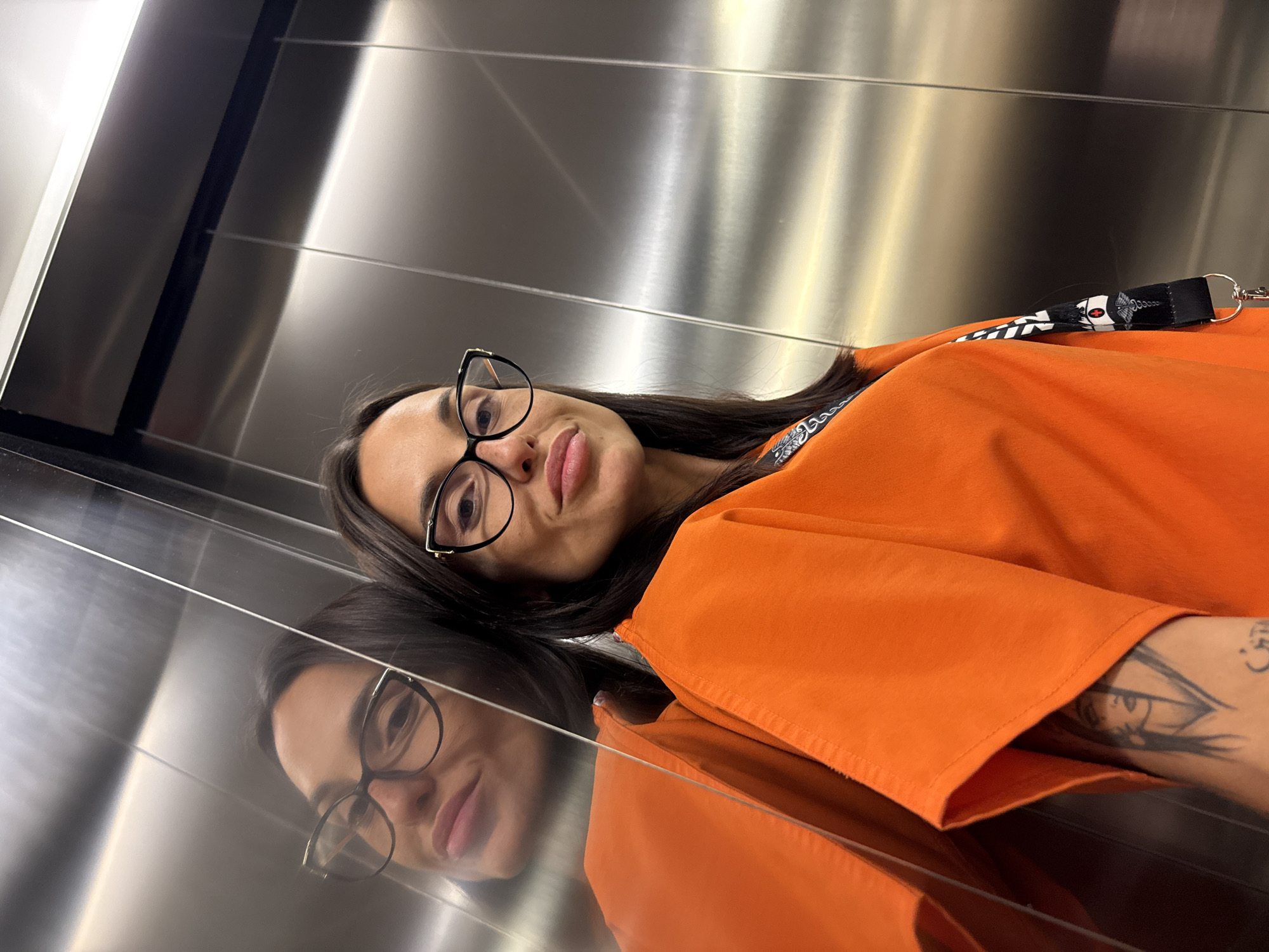 A young woman with dark hair and glasses, wearing an orange top, poses for a selfie in an elevator. The elevator has brushed metal walls that reflect her image. A tattoo of a woman's face is visible on her arm. A lanyard with medical symbols hangs around her neck. The location is the city of Pleven, Bulgaria.