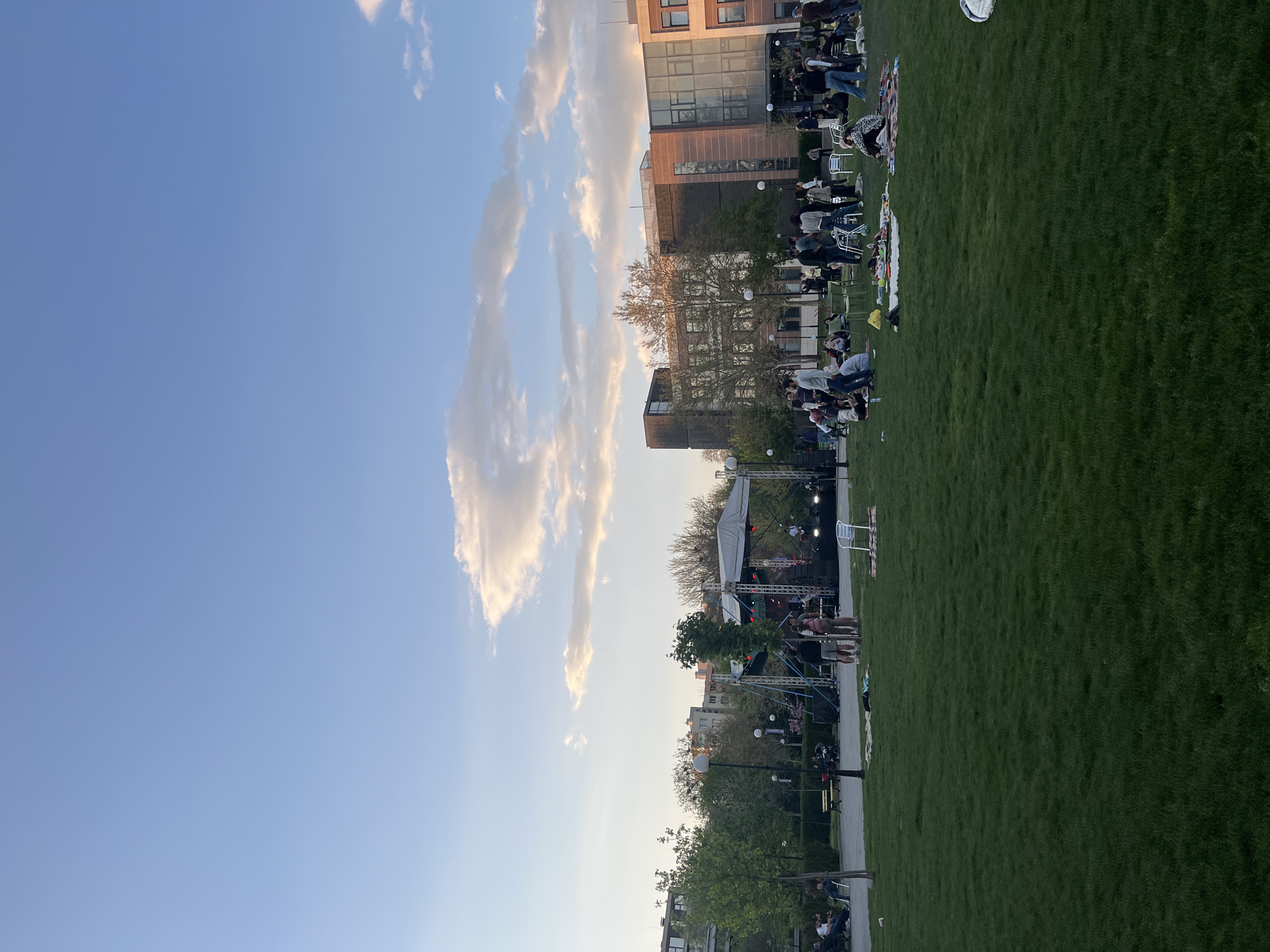 The image depicts a public park in Blagoevgrad, Bulgaria, on what appears to be a late afternoon. A large grassy area hosts a gathering of people seated on chairs and blankets, oriented towards a stage set up with lighting and sound equipment, suggesting a concert or performance event. Buildings surround the park, some featuring modern architectural designs. A clear blue sky with cumulus clouds provides a backdrop. Street lamps line a pathway cutting through the park, indicating pedestrian access. People are interacting with each other, possibly waiting for the event to begin.