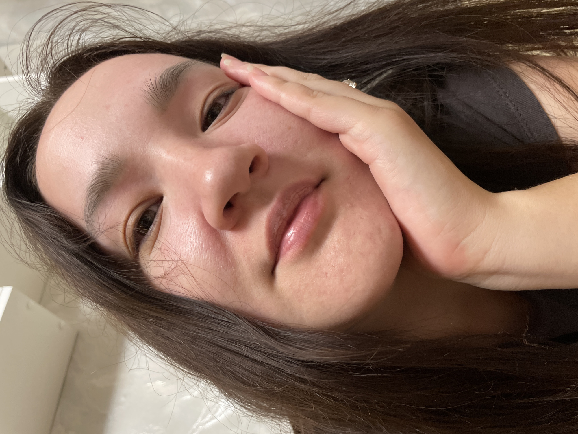 The media file is a close-up, overhead shot of a person's face, tilted. The person has long, dark brown hair that is spread around their face and shoulders. Their skin appears smooth and slightly flushed. They are wearing a black shirt. One hand is placed on their cheek, with fingers extended towards their eye. A ring is visible on the ring finger. The background is out of focus and appears to be a light-colored wall or surface. The lighting suggests it is daytime, possibly indoors. The overall mood is relaxed and contemplative. No text is visible in the media file. The location context provided is Laishevo, Russia.