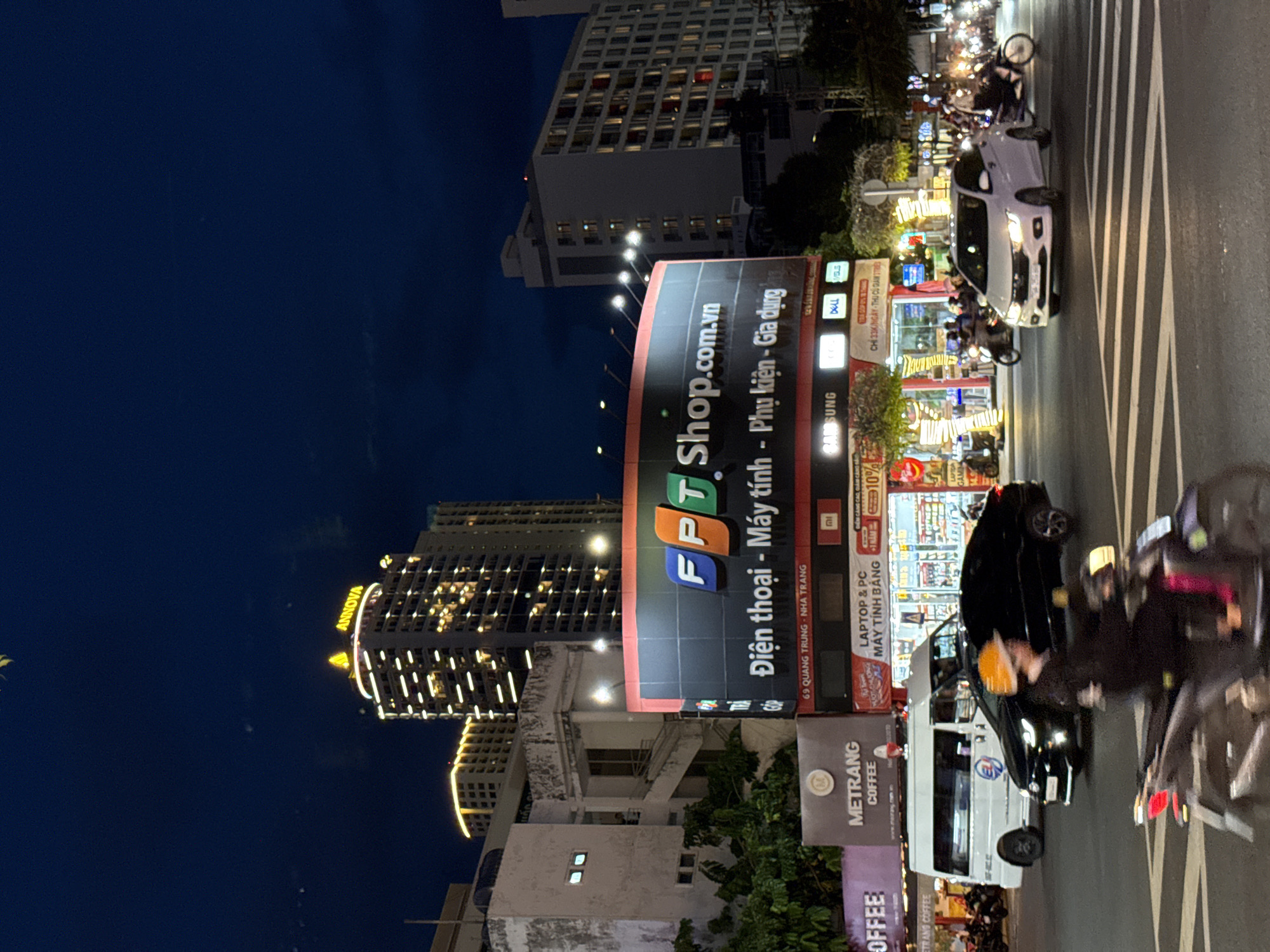 The image depicts a nighttime street scene in Nha Trang, Vietnam. A large, illuminated billboard for "FPT Shop.com.vn" is prominently displayed, advertising "Điện thoại - Máy tính - Phụ kiện - Gia dụng" (phones, computers, accessories, home appliances). Below the FPT billboard, another sign indicates "LAPTOP & PC MÁY TÍNH BẢNG" (laptops & PCs, tablets). Adjacent to the FPT billboard is a sign for "METRANG COFFEE".
The street is bustling with activity, featuring several vehicles including a white van, a black car, a white sedan, and multiple motorcycles. Pedestrians are visible, some on motorcycles and others possibly walking, though their details are blurred due to motion or distance. The street is illuminated by streetlights and the bright lights from the businesses and vehicles. In the background, tall buildings, including a skyscraper with the "ANNOVA" logo illuminated at the top, are visible, indicating an urban environment. The sky is dark blue, suggesting it is either late evening or early morning. The overall impression is of a vibrant, commercial area in a Vietnamese city at night.