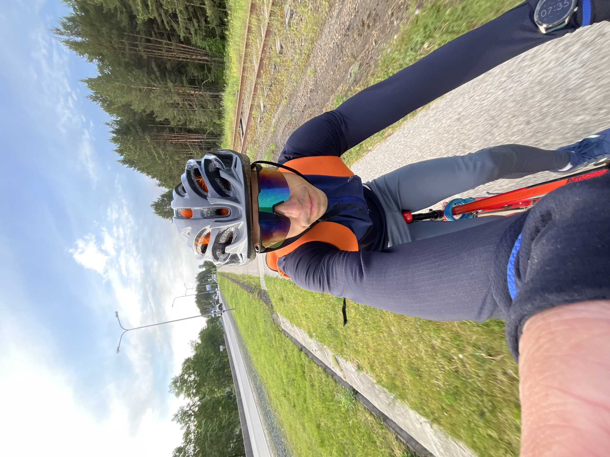 A cyclist is captured in a selfie-style photo, wearing a gray helmet with orange accents, reflective sunglasses, a navy long-sleeved top with orange panels, and gray leggings. The cyclist is riding on a paved path that parallels railway tracks and a grassy area. The background shows a line of trees and the sky. The cyclist is wearing a watch showing the time 07:35. This image was likely taken in the city of Chaikovsky, Russia.