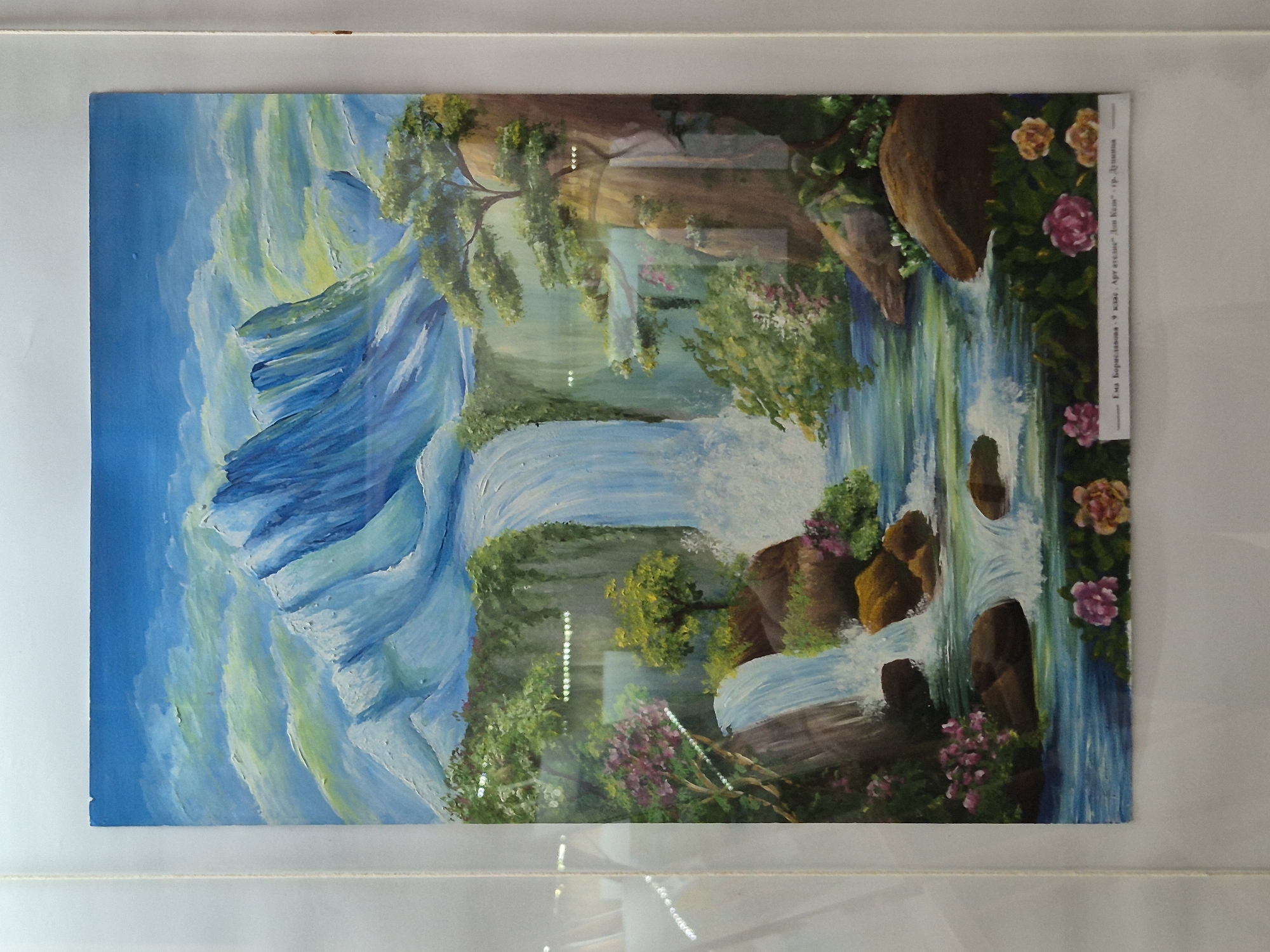 The image shows a painting of a landscape featuring a waterfall cascading down a rocky mountainside. The left side of the painting depicts a large, imposing mountain with blue and white hues, suggesting snow or ice. The waterfall is the central element, flowing from the higher elevations through greenery and trees. Rocks and vegetation are interspersed along the waterfall's path. Flowers bloom on the right side of the painting. The painting is framed and has a paper label attached to the top right corner with Bulgarian Cyrillic text: "Ема Бориславова-9 клас. Ару ателие" Дон Кеш" гр. Дупница". The painting is likely displayed behind glass or plastic, as there are reflections visible on its surface. The location in the city of Srebarna, Bulgaria, is not directly depicted in the image and cannot be inferred from the available visual information.