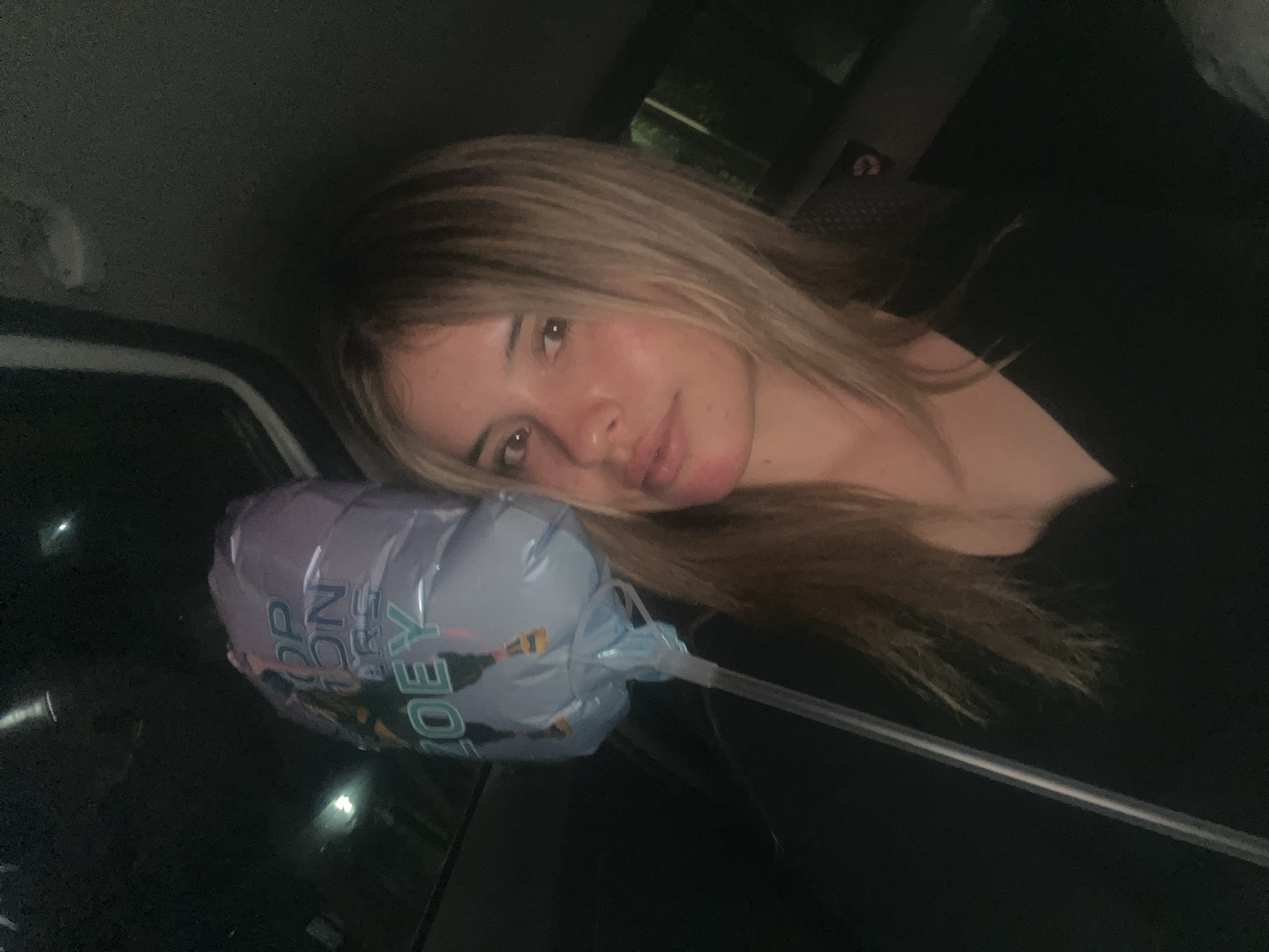 A young woman with blonde hair is lying down in what appears to be the back seat of a car. She is resting her head on a partially deflated balloon that reads "Happy Birthday Joey." The location within San Fernando del Valle de Catamarca, Argentina, is not discernible from the provided image.