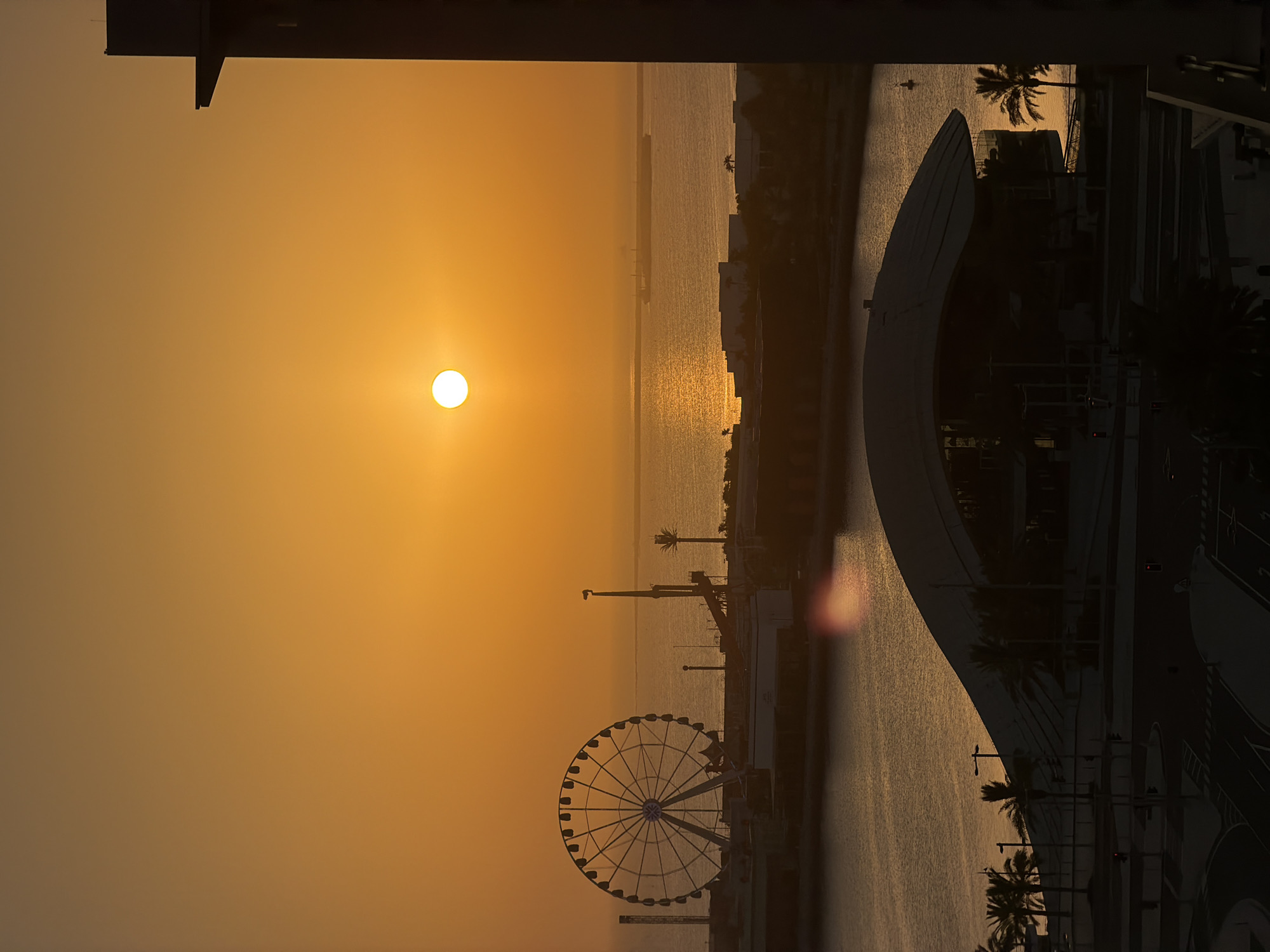 The image depicts a sunset over a waterfront scene. The sun, a bright white orb, hangs in a hazy orange sky, casting a warm glow on everything. The sun's reflection shimmers on the water's surface, creating a golden pathway. In the middle ground, a Ferris wheel stands silhouetted against the sky, its intricate structure a stark contrast to the smooth lines of the buildings and structures that surround it.
Along the waterfront, palm trees are scattered, their fronds appearing dark and wispy in the fading light. Buildings with modern architecture line the shore, some with visible windows and balconies. A curved, futuristic-looking structure dominates a portion of the foreground on the right side of the image. A road with traffic signals is visible in the lower right, suggesting an urban environment.
The overall mood is serene and peaceful, with the warm colors of the sunset creating a sense of tranquility. The scene appears to be taking place in a coastal city or resort area, with the combination of the sea, Ferris wheel, and palm trees. The time of day is clearly sunset, given the position and color of the sun. The weather appears calm, with no visible signs of wind or clouds obscuring the sunset. There is no discernible text in the image.