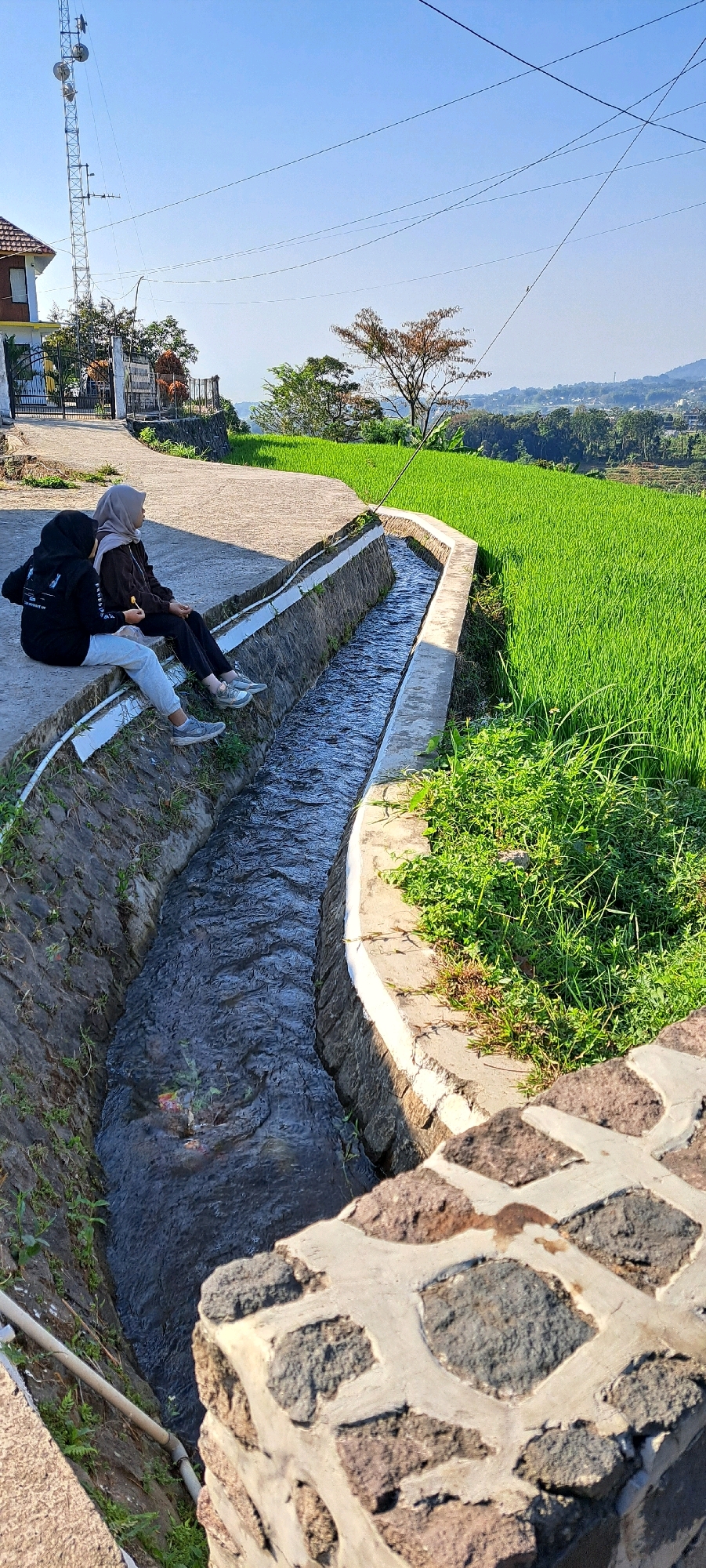 The image shows two young women wearing hijabs, sitting on the edge of a concrete structure that appears to be a man-made irrigation canal. The canal is filled with flowing water and is bordered by stone and grass. The women are facing away from the camera, looking towards a vibrant green rice paddy field. One woman on the left is wearing a black hooded sweatshirt and light-colored pants, with a backpack slung over her shoulder. The woman on the right is wearing a brown sweatshirt and dark pants. The scene is outdoors, with a clear blue sky and a distant view of hills and a small town. A telecommunications tower is visible in the background on the left. The setting is consistent with the rural landscape of Sukoreno, Indonesia, known for its agriculture, particularly rice cultivation. The presence of the irrigation canal suggests a system for watering the fields. The overall impression is one of a peaceful, sunny day in the countryside.