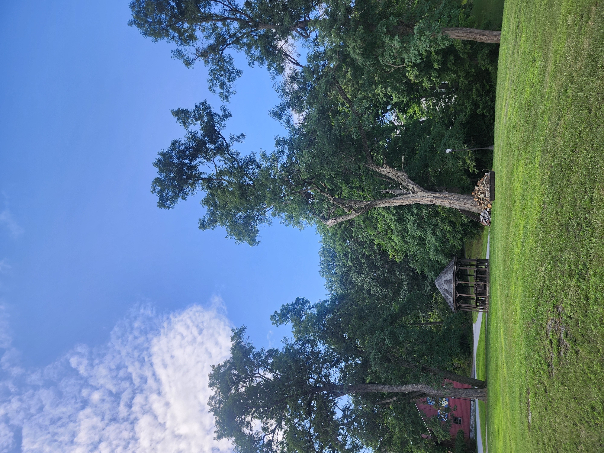 A daytime, eye-level, medium outdoor shot captures a park scene in the Town of Hoosick, United States of America. The sky is bright blue with scattered white clouds. In the foreground, a lush green lawn slopes gently upward. To the left of the lawn, a red building with white trim and stained-glass windows is visible behind a large tree. In the middle ground, a wooden gazebo with a hexagonal roof stands on a paved pathway. To the right of the gazebo, a pile of firewood sits near a lamppost. Tall, mature trees with green foliage dominate the background, creating a dense forest canopy.