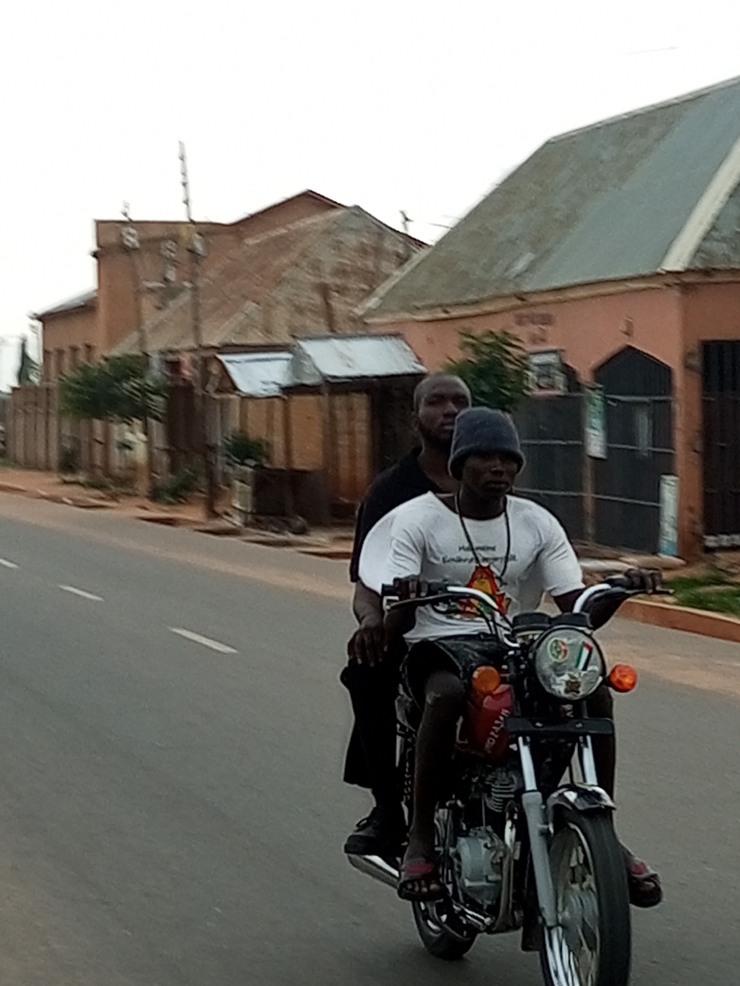 Two individuals are riding on a motorcycle on a paved road in what appears to be the city of Tempure, Nigeria. The person in the front is operating the motorcycle, wearing a dark beanie, a white t-shirt with a graphic design, and dark shorts. The person in the back is wearing a black shirt and appears to be looking forward. The motorcycle is red and black, with chrome accents. In the background, there are several buildings with reddish-brown brick and corrugated metal roofs, typical of urban architecture in the region. Vegetation, including trees and bushes, is visible along the roadside. The sky is overcast, suggesting a dull or hazy day. The road has white lane markings.