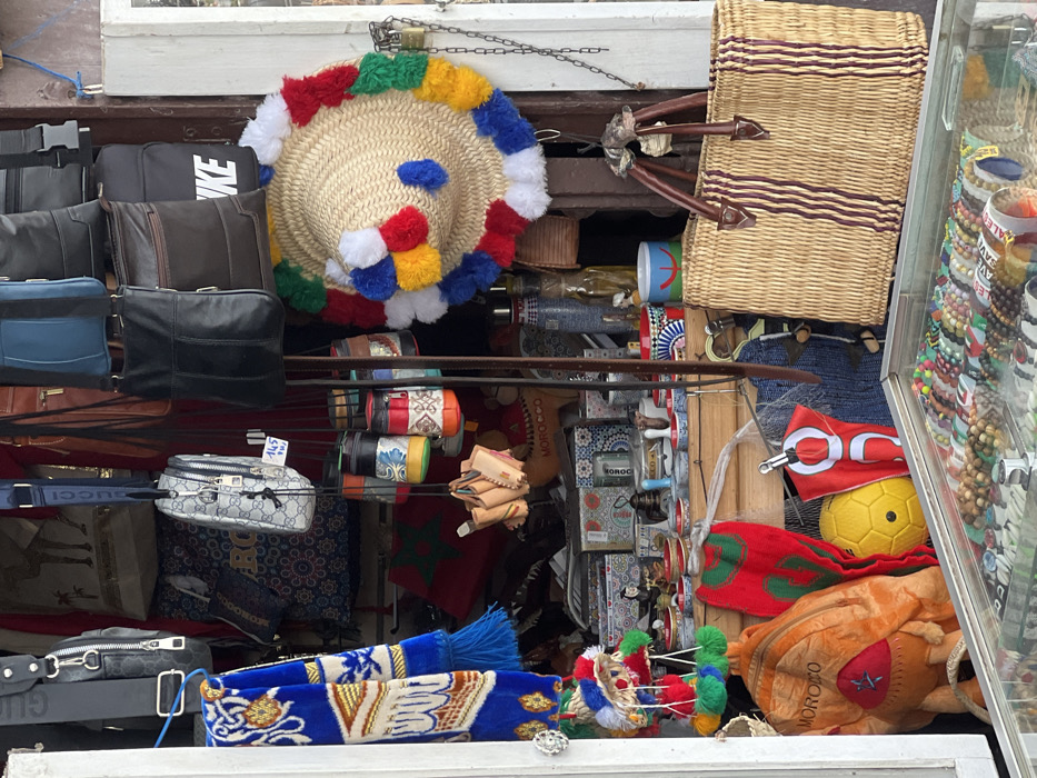 The image displays a densely stocked souvenir shop entrance in Tanger, Morocco. No individuals are visible. The shop's opening is framed by white jambs on the left, revealing a cluttered interior and hanging merchandise.
In the upper section, various handbags and messenger bags are suspended, including dark leather models, a light gray patterned bag with a "GUCCI" branded strap, and a black bag partially obscuring a "NIKE" logo. A small patterned bag displays a tag with "145 DH." Below these, a cluster of small, cylindrical containers with colorful patterns and several miniature light brown leather objects hang.
A prominent, round straw hat adorned with numerous colorful pom-poms (red, blue, yellow, green, white) hangs to the right of the shop's center. To its lower right, a large woven straw tote bag with brown leather handles and dark vertical stripes is suspended.
The lower foreground features a glass display case containing multiple rows of bracelets and necklaces crafted from beads and various materials. Further inside the shop, visible behind the hanging items, are shelves stacked with packaged goods, small ceramic bowls, tea glasses, glass bottles, and a visible red Moroccan flag. Additional items on display include an orange backpack with "MOROCCO" text, a yellow soccer ball, and smaller woven hats with pom-poms.