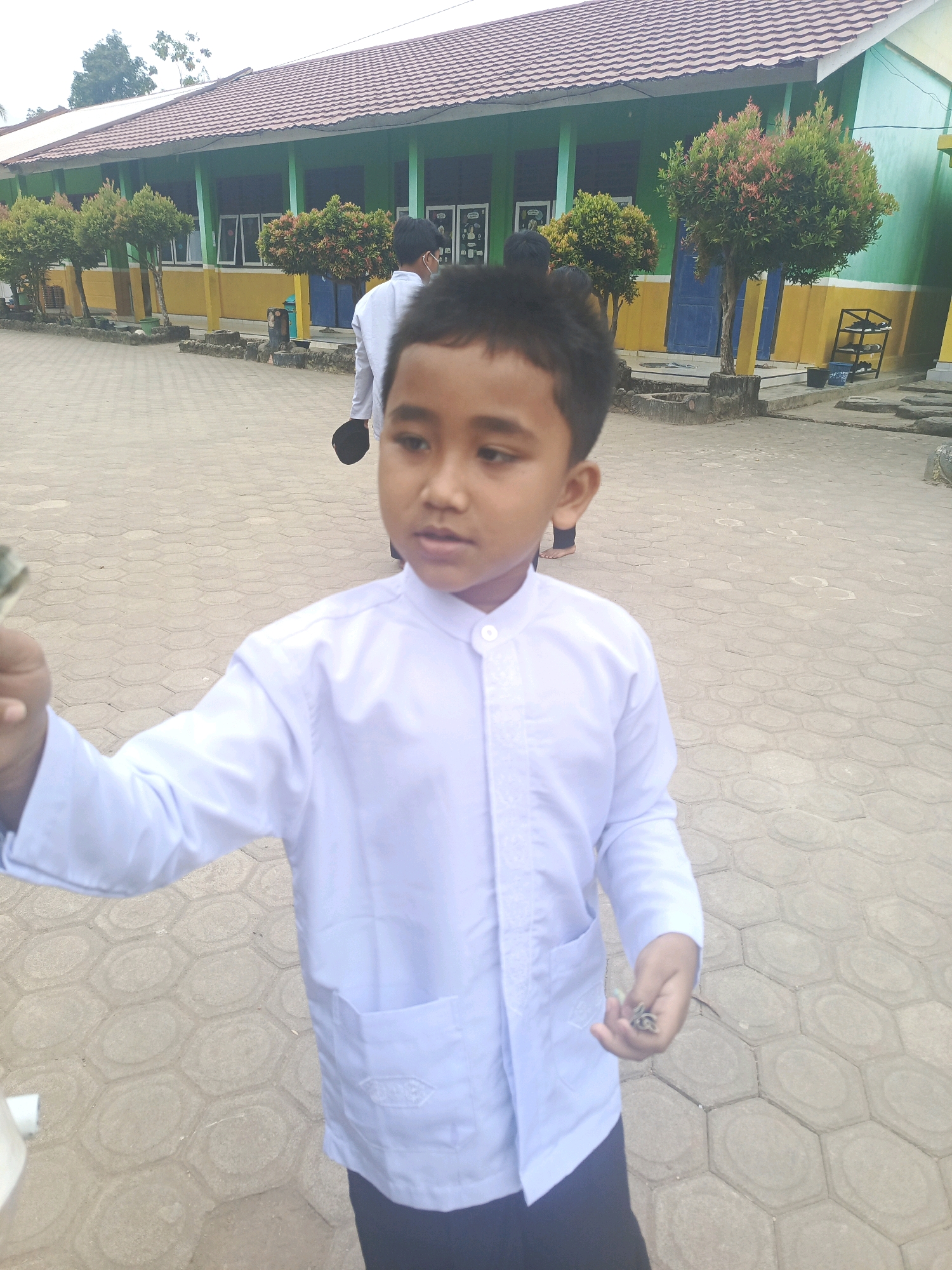 A young boy wearing a white school uniform stands in a paved schoolyard in Megang Sakti II, Indonesia. He is holding some small objects in his right hand and appears to be looking at them. Behind him, a building with a green facade and a red tiled roof is visible, lined with decorative trees. Another student in a similar uniform is walking away from the camera in the background. The ground is covered with hexagonal paving stones.