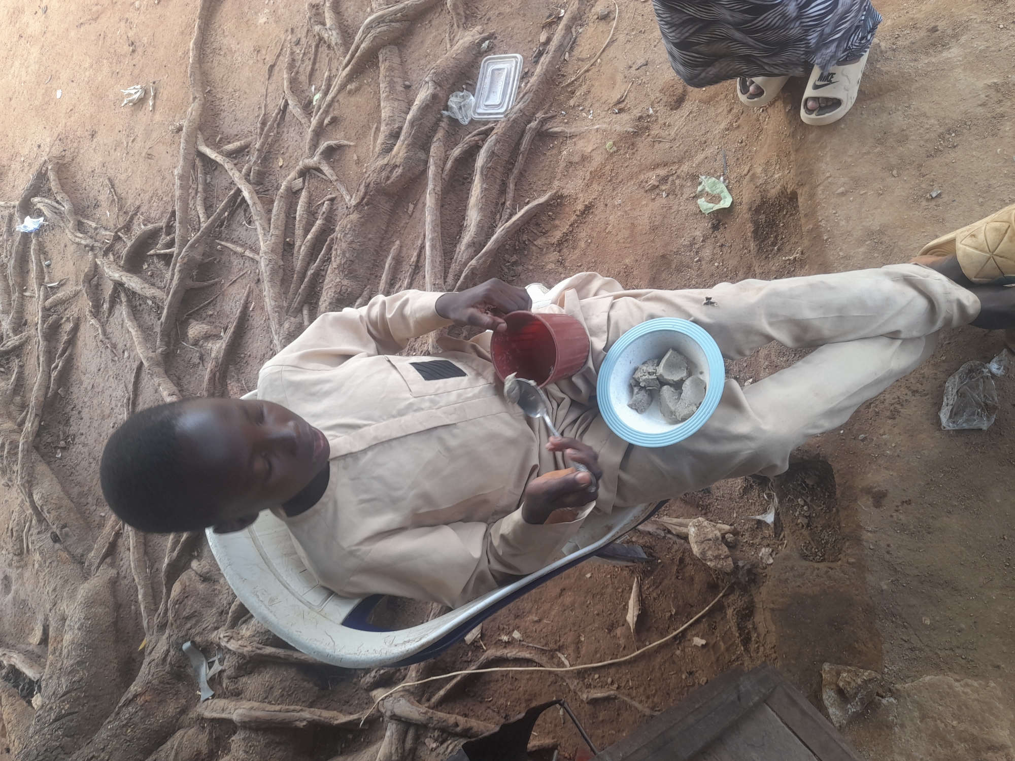 Akwanga, Nigeria: An individual wearing a light-colored uniform sits on a white plastic chair on bare soil next to exposed tree roots. The person holds a spoon in one hand and a red cup in the other. A light blue bowl containing gray, irregularly shaped objects rests on their lap. Nearby, someone wearing black and white patterned clothing stands with white Nike sandals. The ground is visible and contains dirt, roots and debris.