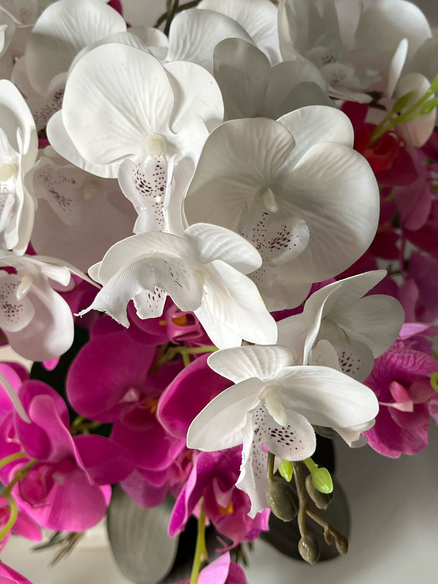 This is a close-up, overhead, vertical shot of a bouquet of artificial orchids in Tungkaran Pangeran, Indonesia. The bouquet features a mix of white and vibrant pink orchids. The white orchids have delicate dark speckles on their petals, while the pink orchids are a solid, bright hue. Some green buds are visible on a stem in the lower right portion of the frame. The background is blurred and appears to be a neutral gray. The lighting is soft and diffused, highlighting the textures of the artificial petals. No people or animals are present in the image.