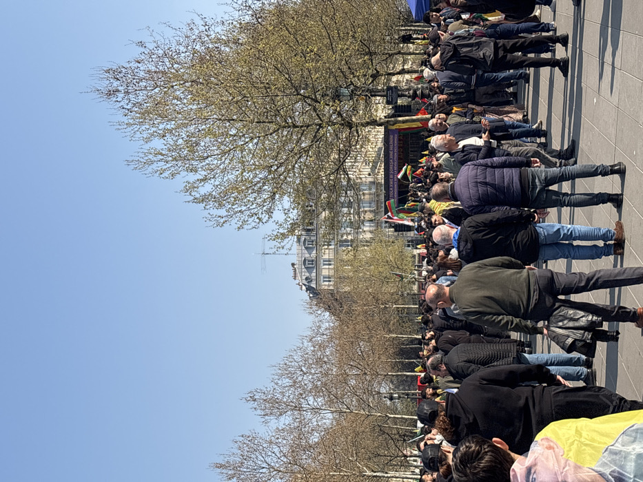 A wide-angle, eye-level photograph captures a dense outdoor gathering of several hundred individuals under a clear blue sky on a sunny day in what appears to be Place de la République, Paris, France.
The foreground is filled with the backs and sides of numerous people, predominantly adult males dressed in casual dark clothing, including jackets, hoodies, and jeans. They stand on a paved surface, facing towards the middle ground.
In the middle ground, a large crowd extends backward towards a stage or platform. Visible within the crowd and near the platform are multiple flags, characterized by red, white, and green horizontal stripes with a yellow sun emblem in the center, identifiable as Kurdish flags. A prominent purple banner is mounted on the stage, displaying white text in Kurdish: "TOLA XWE Bİ ŞOREŞA JÎNE JÎLÎNÎ! DENDÊRATA XWE Bİ REVOLUSTYONA JINÊ!" with a smaller line in French below: "DÉFENDONS LA RÉVOLUTION FÉMINISTE!" The crowd appears to be observing or listening to the activity at the stage.
The background features rows of deciduous trees, many with nascent green leaves or still bare branches, indicating early spring. Beyond the trees, classic multi-story Parisian buildings with light-colored stone facades and mansard roofs are visible, confirming an urban setting in Paris. The overall scene depicts a large, peaceful public assembly or protest.