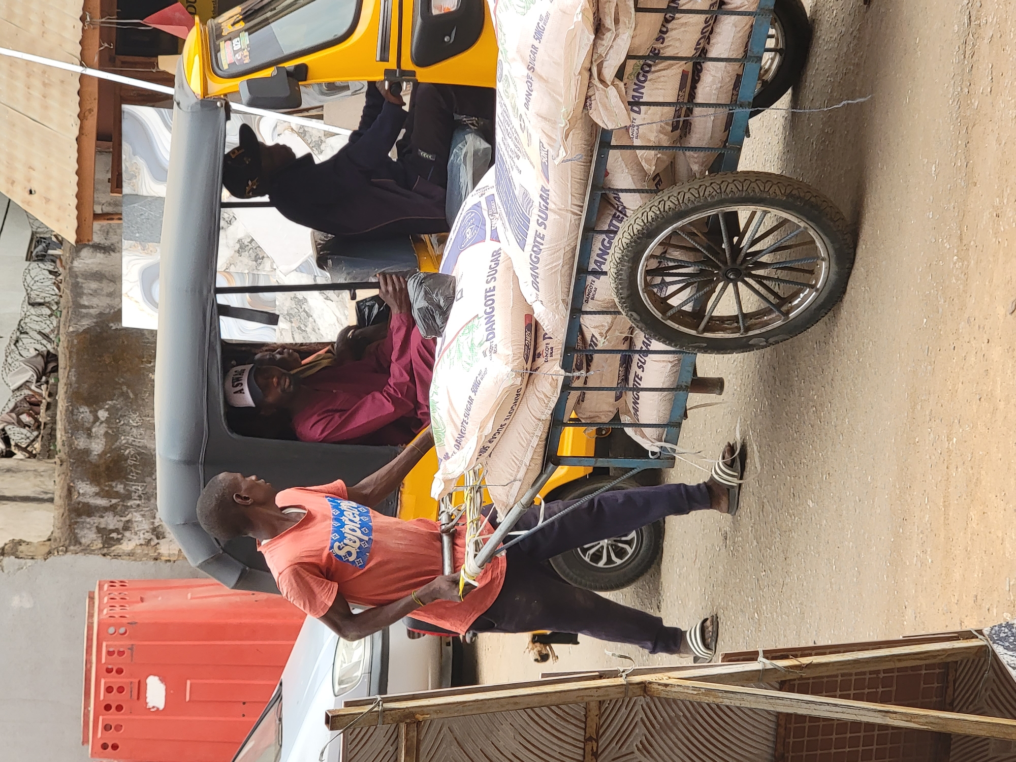 The image depicts a man pulling a cart laden with bags of "DANGOTE SUGAR 50KG". The cart is attached to a yellow, three-wheeled motor vehicle commonly known as a "Keke Napep," which is occupied by at least three individuals. The man pulling the cart is wearing an orange t-shirt with blue lettering, dark trousers, and sandals. The environment appears to be an urban street, with buildings and other vehicles visible in the background. The OCR data suggests the brand of sugar and its weight. The location may be Maiburiji, Nigeria.