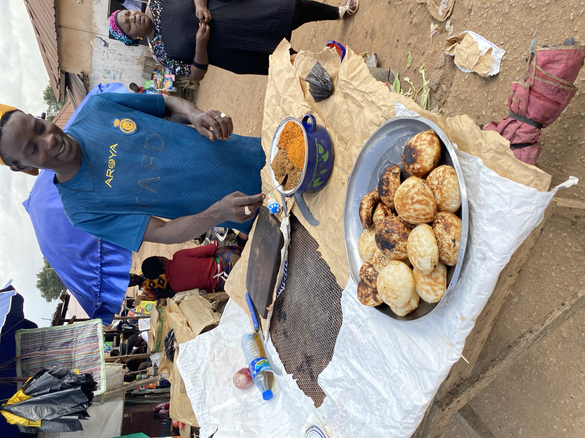 In Bukuru, Nigeria, a street vendor displays a plate of golden-brown, round pastries. The pastries are arranged in a metallic dish, piled high on a stall covered with brown paper and white plastic sheeting. Beside the pastries, a blue pot holds two distinct piles of spices, one appearing reddish-orange and the other a lighter brown. A man, likely the vendor, wears a blue shirt with yellow lettering and a yellow cap, and is seen reaching towards the spices. To his right, a woman in a black dress and colorful headscarf observes the scene. The background shows a bustling market environment with umbrellas, stalls, and buildings with corrugated iron roofs, suggesting a typical day in the area.