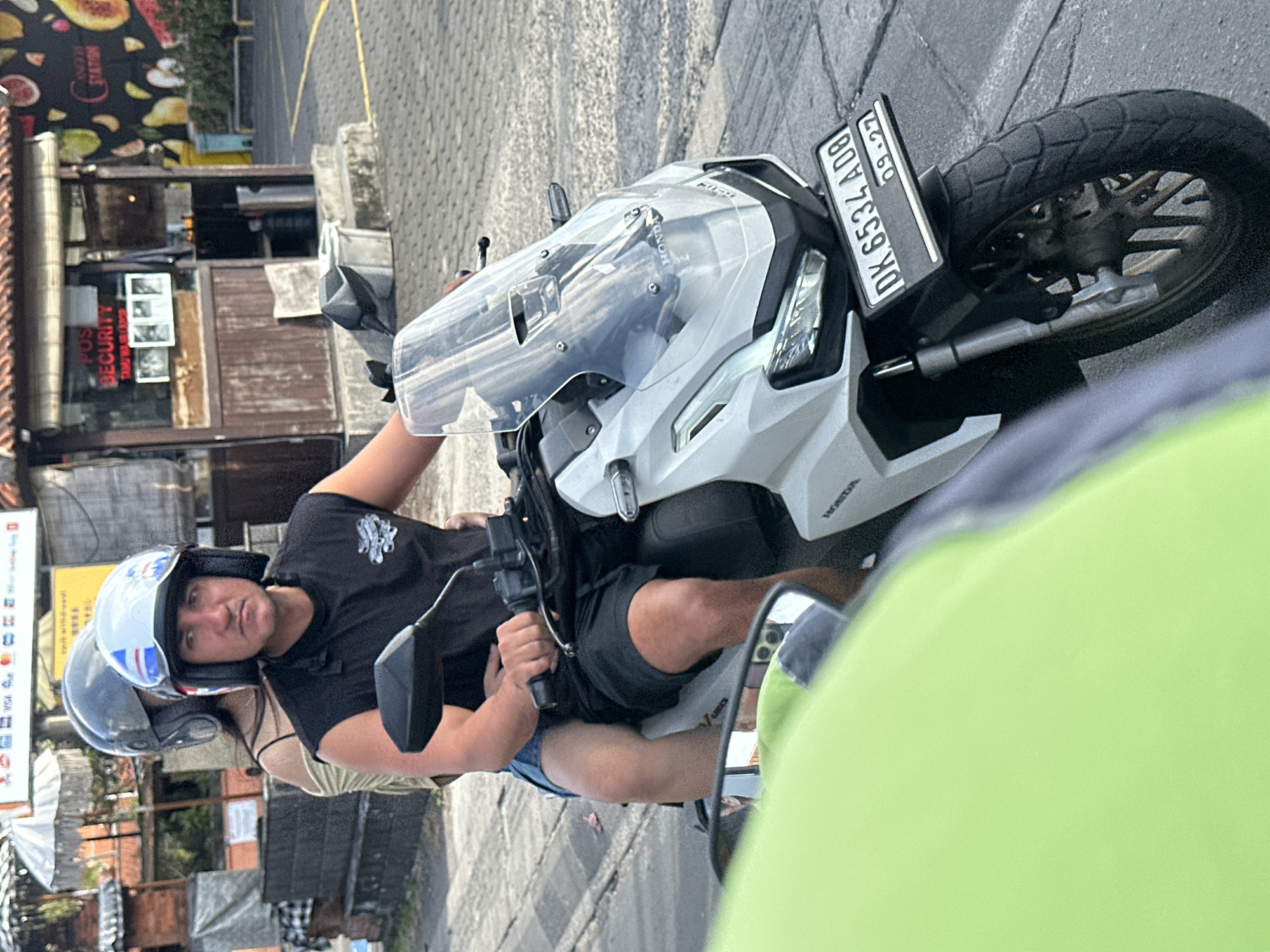 An adult male, wearing a black sleeveless shirt and black shorts, rides a white Honda scooter. He is wearing a white helmet with red, white, and blue stripes. An adult female, wearing a beige tank top and denim shorts, is a passenger on the scooter. She is wearing a grey helmet. The scooter has a black license plate with "DK 6534 A08" and "09.27" printed on it. The scooter is on a paved road, with a sidewalk and buildings in the background. A light green object, possibly another vehicle, is in the foreground, partially obscuring the view of the scooter. The scene appears to be in a city, likely North Kuta, Indonesia, based on the license plate format. A small security booth is visible to the right of the scooter.