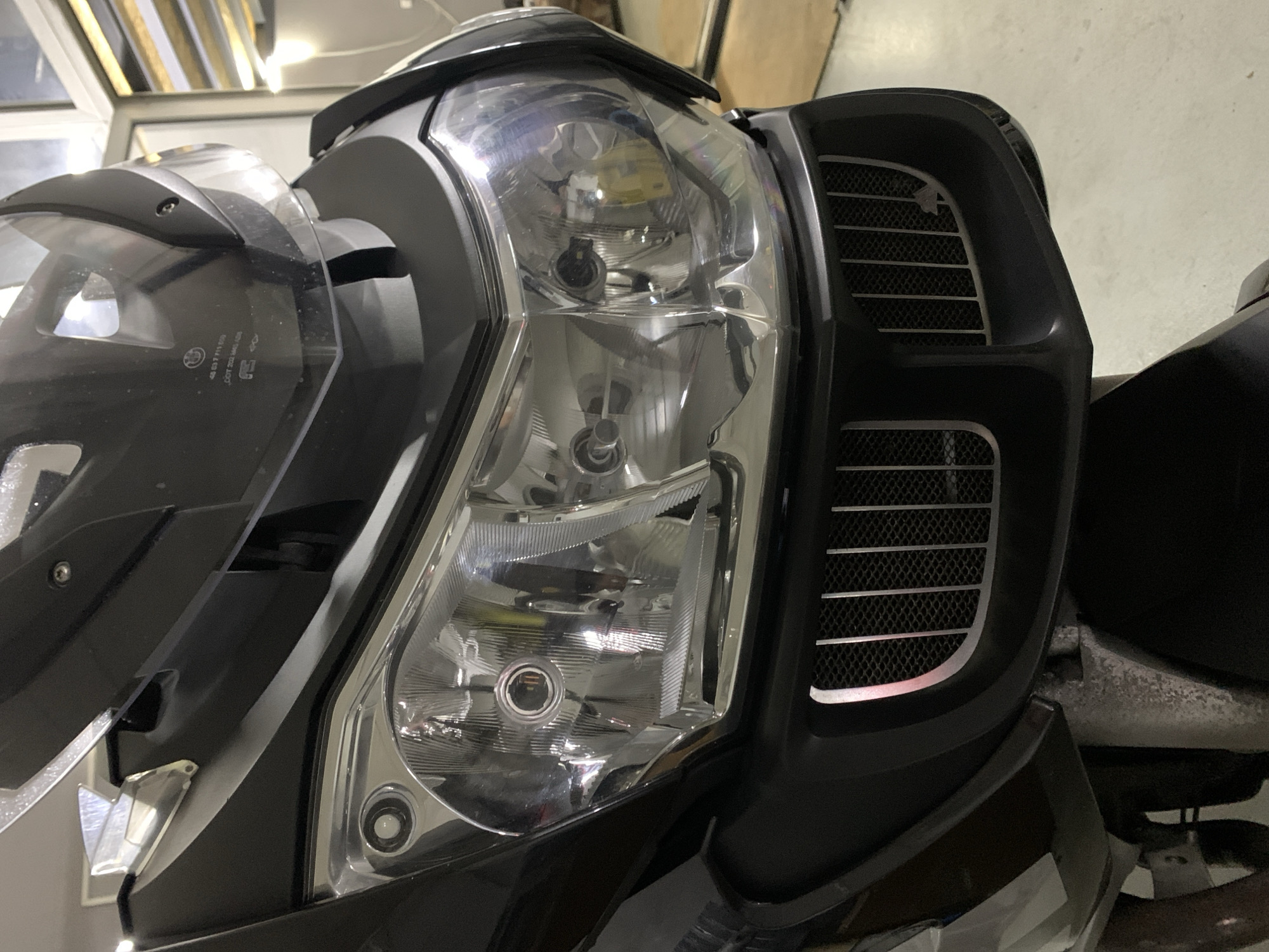 The image shows a close-up view of the front of a black motorcycle, focusing on the headlights and the front grille. The motorcycle appears to be stationary, possibly in a garage or workshop, as indicated by the muted lighting and what appears to be a concrete floor in the background. The headlights are prominent, with clear lenses and visible internal reflectors. The front grille is a dark, textured material with silver vertical slats. There are no people visible in the image, and no specific environmental details that would allow for pinpointing the exact location in Kobryn, Belarus. The scene is static and objective, with no discernible action or interaction occurring.