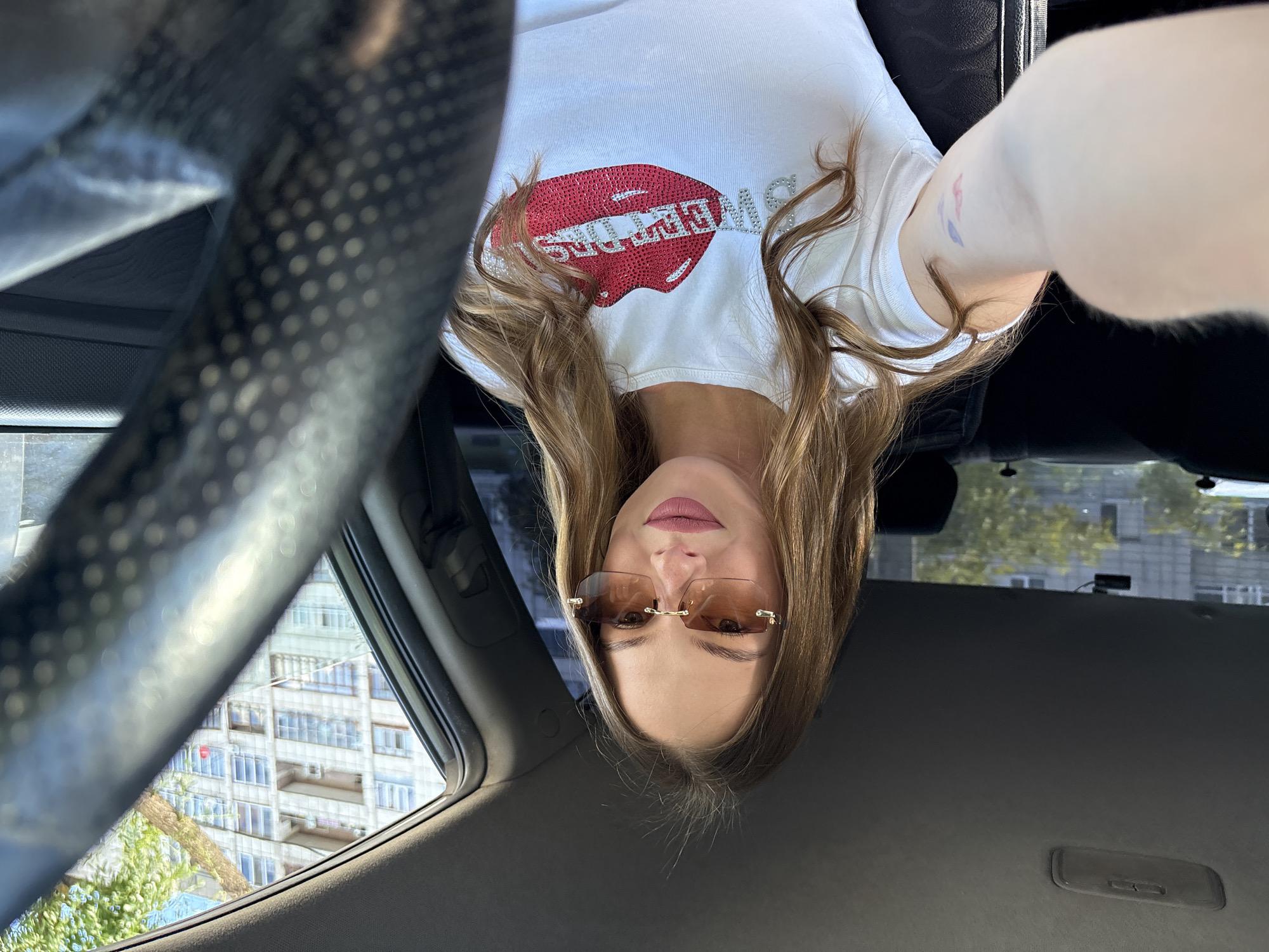 A young woman with long, wavy brown hair and light brown eyes is sitting in the driver's seat of a car, taking a selfie. She is wearing oversized, rimless sunglasses with a light brown tint and a white t-shirt with a large red sequined graphic of lips and the word "SWEETNESS" spelled out in rhinestones. The car's interior is visible, including the dashboard, steering wheel, and the back seats. Outside the car, a multi-story building with many windows and balconies, along with some trees, can be seen. The scene appears to be set on a sunny day in a city, possibly Perm, Russia, given the architectural style of the building.