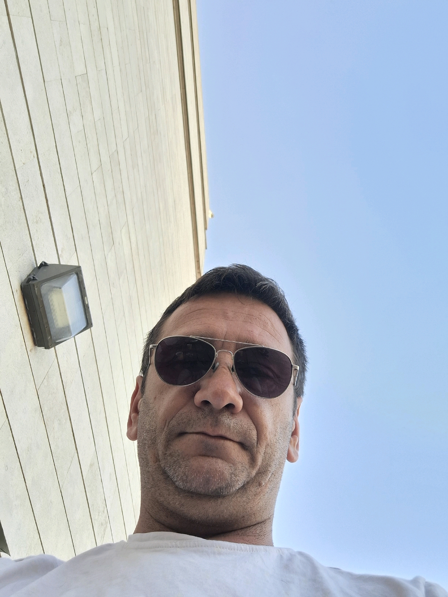 The image is a selfie taken from a low angle, looking up at a man. He is wearing sunglasses and a white t-shirt. The background consists of a light-colored, textured wall and a clear blue sky. An outdoor light fixture is attached to the wall to the left of the man. The scene appears to be outdoors in an urban environment. There is no indication of the specific location within Kiryat Gat, Israel, beyond it being an outdoor setting.