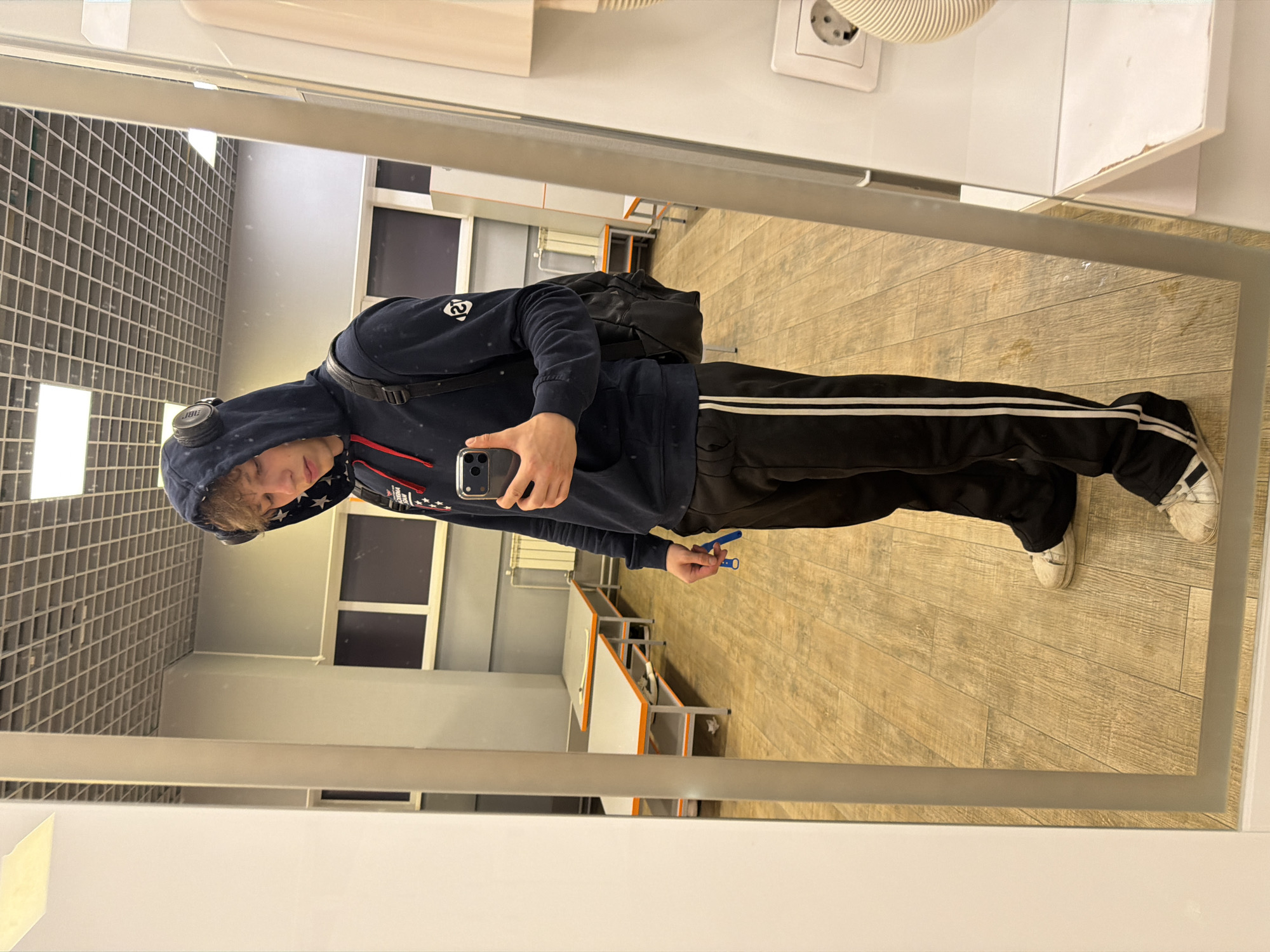 The image is a mirror selfie taken by a young male in what appears to be a locker room or changing area. The individual is wearing a dark blue hooded sweatshirt with red drawstrings, black track pants with white stripes, and white athletic shoes. He has headphones on and is holding a smartphone in his right hand, taking the picture. A black backpack is slung over his left shoulder. In the background, visible in the mirror's reflection, are several tables with orange trim, a wall with a radiator, and a tiled ceiling. The floor is made of light-colored wood planks. The overall environment suggests a utilitarian space, possibly part of a gym, school, or sports facility. There is no specific visual information to ascertain the exact location within Nizhny Novgorod, Russia.