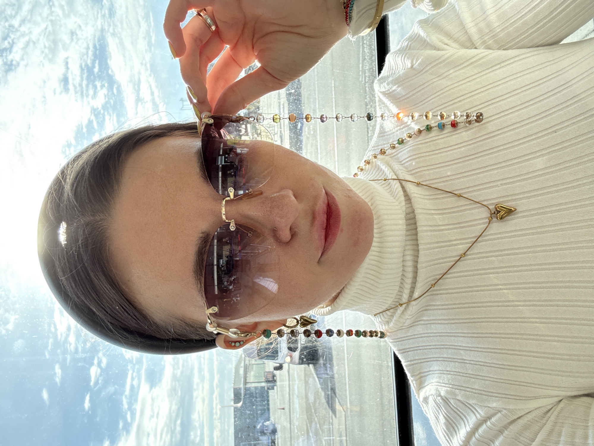 A young woman with dark hair is holding her sunglasses up with her right hand, and wearing a white turtleneck sweater. She is seated by a window with a view of Khimki, Russia, based on the reflected scenery. The sunglasses have a gold frame, and are attached to a beaded chain. She is also wearing a gold necklace with a heart pendant, and multiple rings.