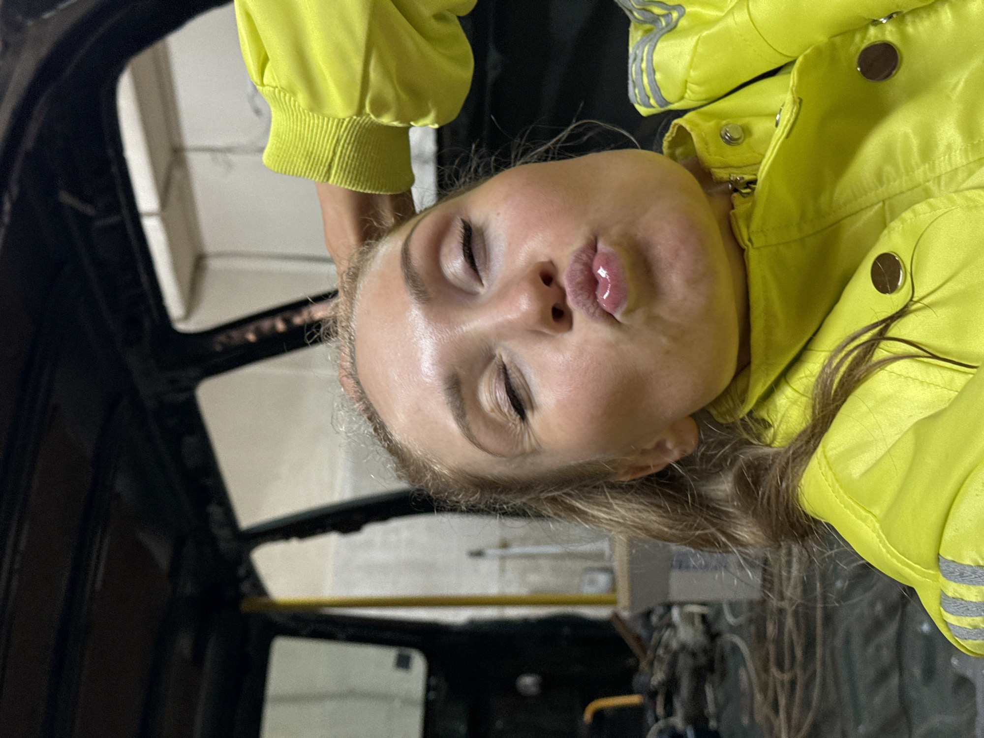 A woman with her eyes closed and lips puckered is partially visible inside what appears to be the stripped interior of a vehicle. She is wearing a bright yellow jacket with reflective stripes. The interior of the vehicle is black with visible metal framing and part of the windshield. The setting suggests an industrial or maintenance location. There is no verifiable information to confidently place this scene within the city of Perm, Russia.