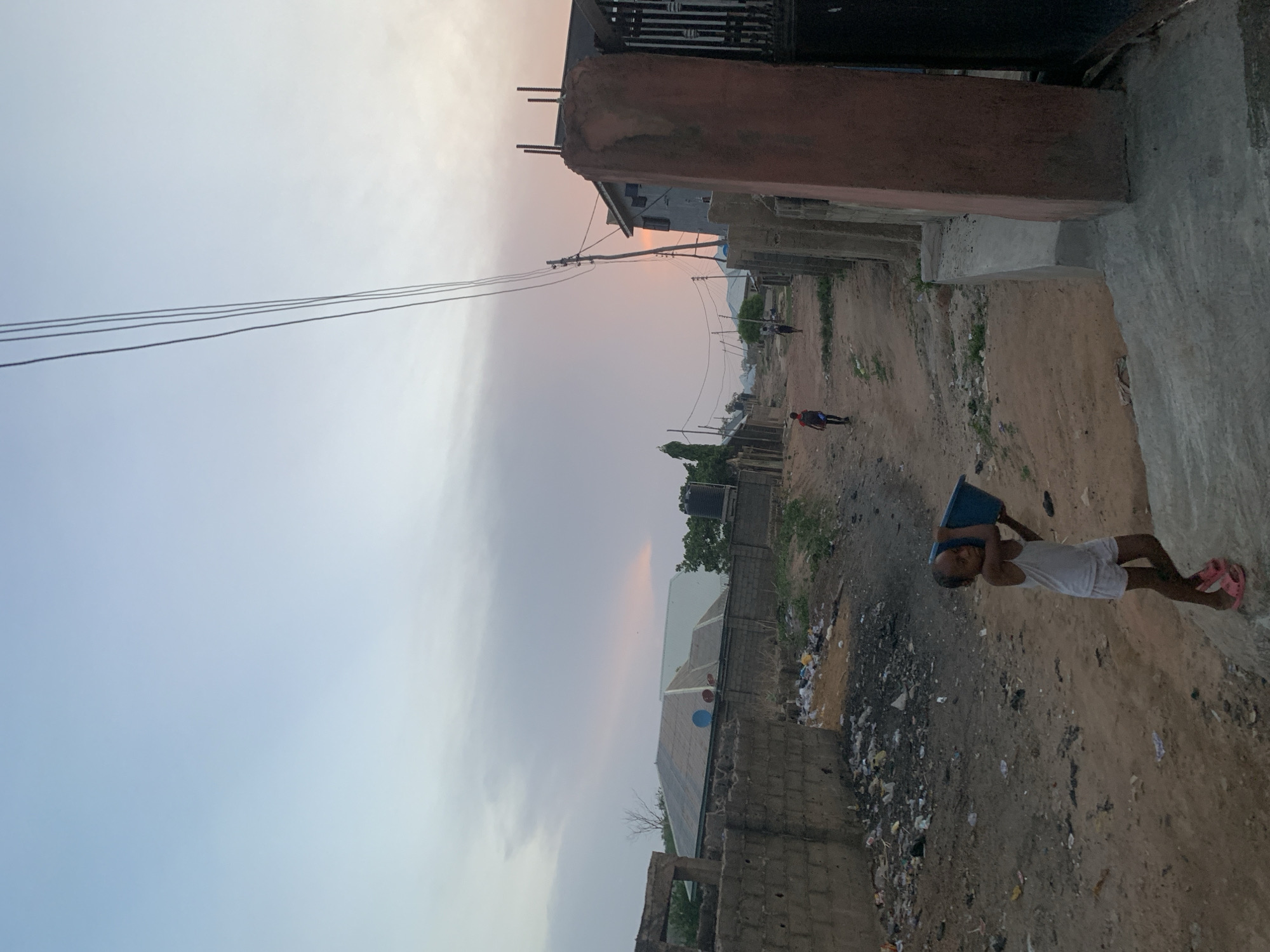 The image depicts an outdoor scene in Lokoja, Nigeria. The sky is overcast with a hint of sunset. A young child dressed in a white top and shorts, and pink sandals is walking along a dirt road carrying a blue rectangular container on their head. Other people can be seen in the distance. The surrounding environment includes buildings in varying states of construction, overhead electrical wires, piles of trash, and some vegetation. The location appears to be a residential area with unpaved roads.
