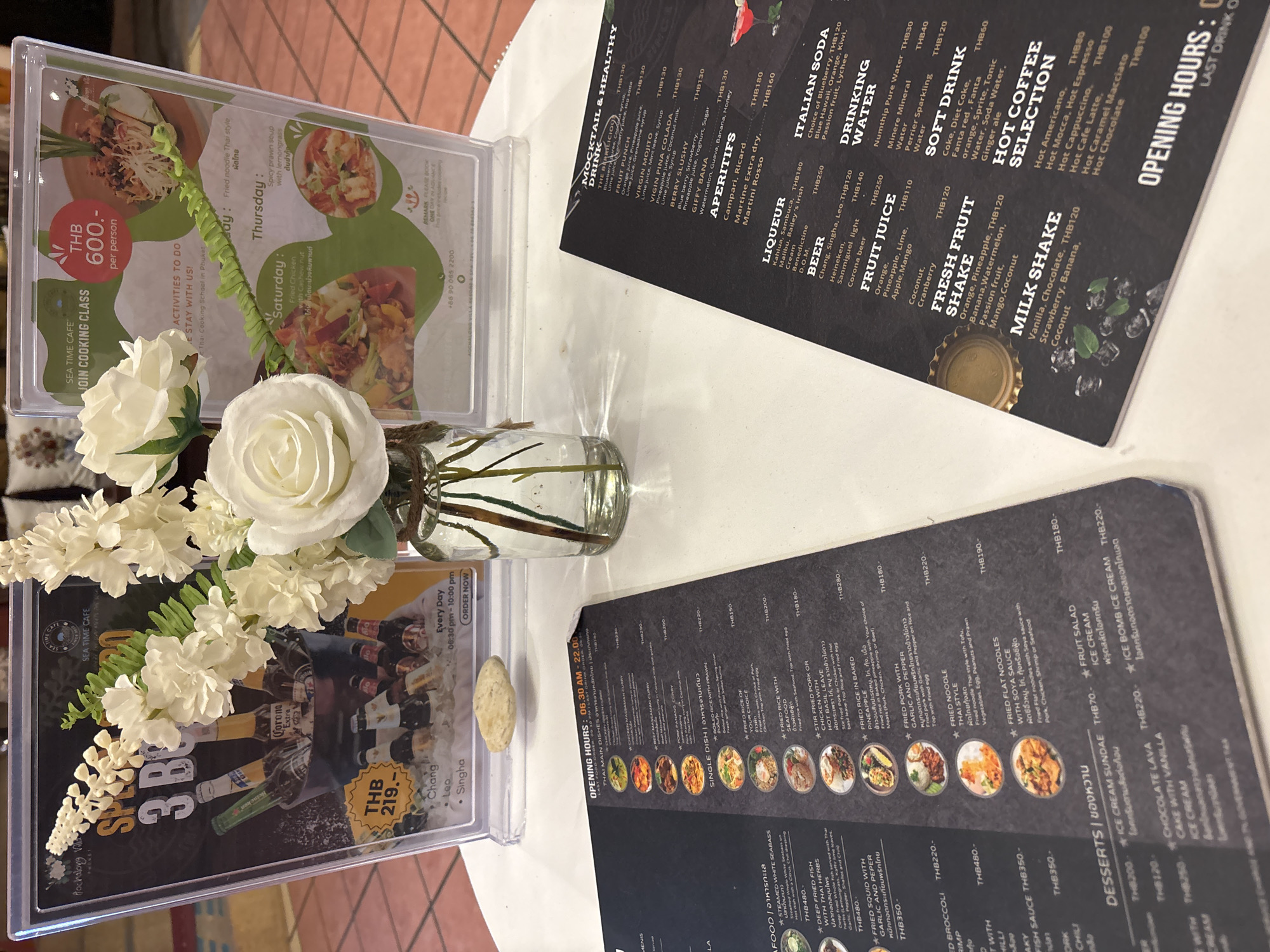 The image shows a table at a cafe, likely in Chalong, Thailand, with a menu and decorative flowers. In the foreground, a dark menu lists various food and drink items, including "Thai Main Dishes," "Single Dish," "Desserts," "Mocktail & Healthy," "Aperitifs," "Liqueur," "Beer," "Fruit Juice," "Fresh Fruit Shake," "Milk Shake," "Italian Soda," "Drinking Water," "Soft Drink," and "Hot Coffee Selection." The prices are listed in Thai Baht (THB). To the right of the menu, a clear plastic frame displays a promotional advertisement for "SEA TIME CAFE" offering a "JOIN COOKING CLASS" for THB 600 per person. This advertisement also highlights daily activities with specific dishes offered on certain days, such as "Fried noodle Thai style" on Thursday and "Spicy prawn soup with lemongrass" on Friday.
In the upper portion of the image, a bouquet of white flowers, including roses and other delicate blossoms, is arranged in a glass vase, partially obscuring the promotional advertisement. Behind the flowers, a dark sign advertises "3 BEER" for THB 219 and lists beer brands like Chang, Leo, and Singha. The environment suggests an indoor seating area of a cafe or restaurant, with hints of furniture and possibly outdoor elements in the background. The overall scene is clean and well-lit, with a focus on the cafe's offerings and promotional materials. There are no people visible in the image.
