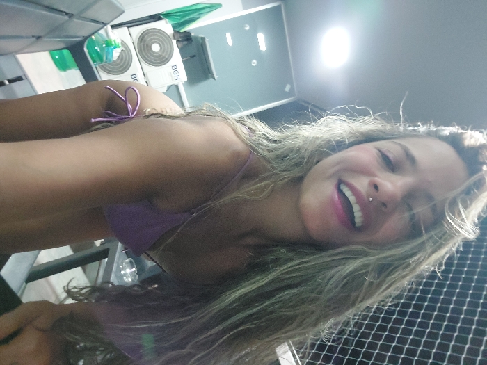 A young female individual with long, wavy blonde hair and a philtrum piercing is depicted smiling broadly, looking directly at the camera. She is wearing a light purple bikini top and matching bottom, and is bending forward as if taking a selfie. The scene is a residential balcony in Buenos Aires, Argentina. Visible to the left of the subject are two stacked outdoor air conditioning units, both bearing the "BGH" brand logo, a manufacturer primarily associated with Argentina. A green towel or fabric hangs nearby, and a metal-slatted chair is partially visible. To the right, a white safety net, characteristic of apartment balconies, covers the upper background. A dark-colored table holds a clear glass ashtray containing a cigarette butt. An overhead circular light fixture is visible on the upper left side of the frame.