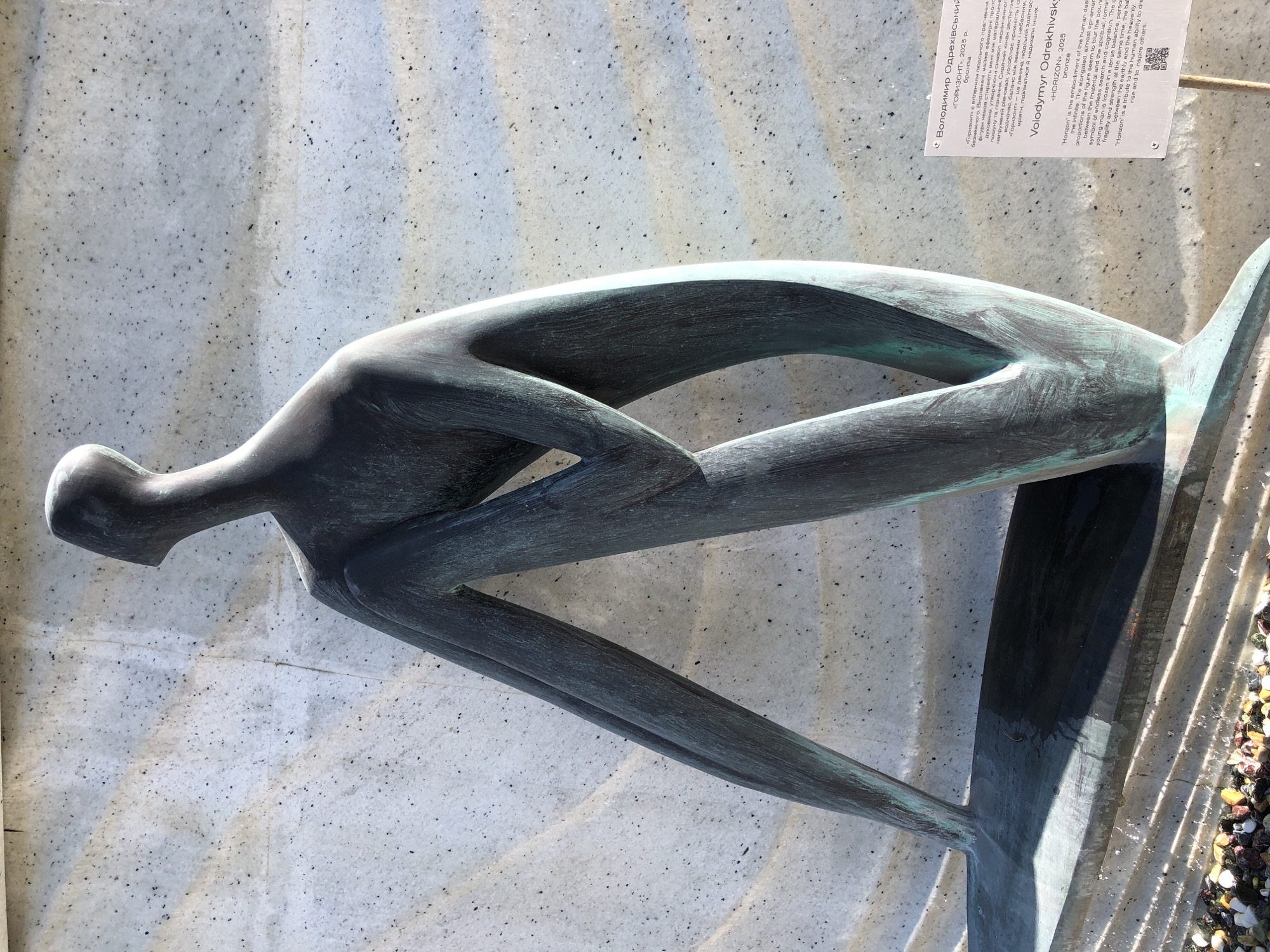 The image shows a bronze sculpture of a stylized human figure in a seated position, with its legs extended forward. The sculpture has a smooth, elongated form and a weathered, greenish-blue patina. It is positioned in front of a light-colored, textured wall with a wavy pattern. To the right of the sculpture, a white information plaque is visible, mounted on a thin pole. The plaque contains text in both Ukrainian and English. Below the sculpture, a collection of colorful pebbles is arranged. The overall scene suggests an outdoor public art installation. The information plaque identifies the sculpture as "HORIZON" by Volodymyr Odrekhivsky, created in 2025. Based on the text on the plaque, this artwork is located in Lviv, Ukraine.