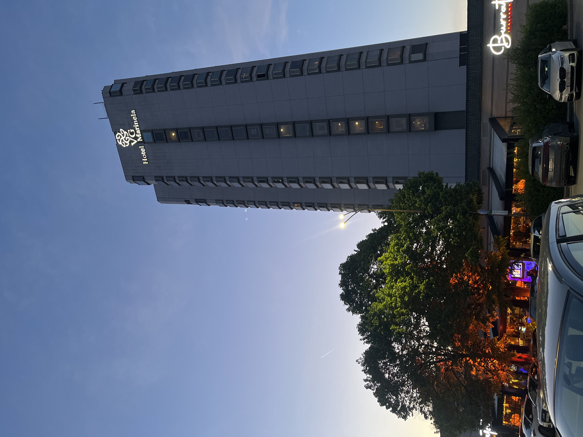The image shows the Hotel Marinela in Sofia, Bulgaria, a tall, modern building with a light gray facade. The hotel's logo is visible near the top. Adjacent to the hotel is a restaurant called Burrata Italiana, illuminated with signage. Several cars are parked in front of the building and restaurant. A large tree is located between the hotel and the restaurant, partially obscuring the view. The sky is a clear, light blue. The lighting suggests it is late afternoon or early evening.