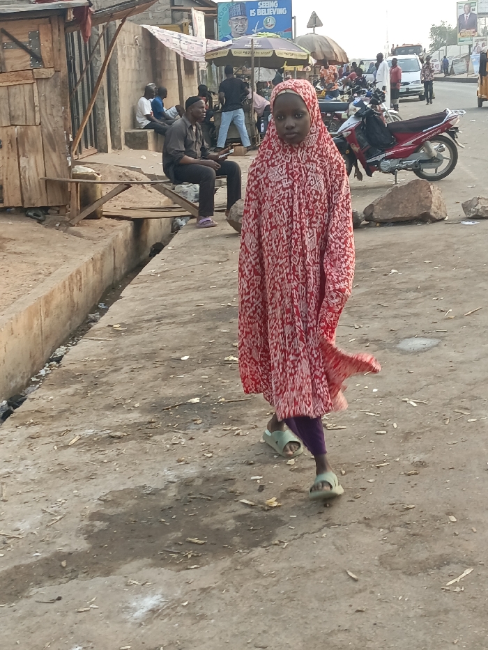 An outdoor scene in Alizaga, Nigeria, depicts a street with pedestrian and vehicle activity. In the immediate foreground, a young person, possibly female, stands or walks toward the viewer. They wear a red and white patterned full-length garment covering the head and body, purple trousers visible beneath the dress, and light green slide sandals. Their gaze is directed forward.
To the left, a concrete sidewalk or raised platform features a shallow open drainage ditch filled with debris. Adjacent to this, crude wooden structures and a concrete wall are visible. A man in a dark shirt, dark trousers, and a dark cap is seated on a makeshift wooden bench alongside the ditch, holding a mobile phone. Further back on the left, other individuals are seated or standing near structures.
The right side of the image shows a paved road. A red motorcycle is parked on the shoulder, with large rocks positioned nearby. In the mid-ground, a triangular street sign is visible. Further down the road, additional vehicles, including cars and trucks, are present, alongside various pedestrians and street vendors operating under umbrellas. A prominent billboard in the background displays text including "SEEING IS BELIEVING" and the image of a political figure. The ground throughout the visible area is primarily concrete, appearing dusty and worn, with scattered debris.