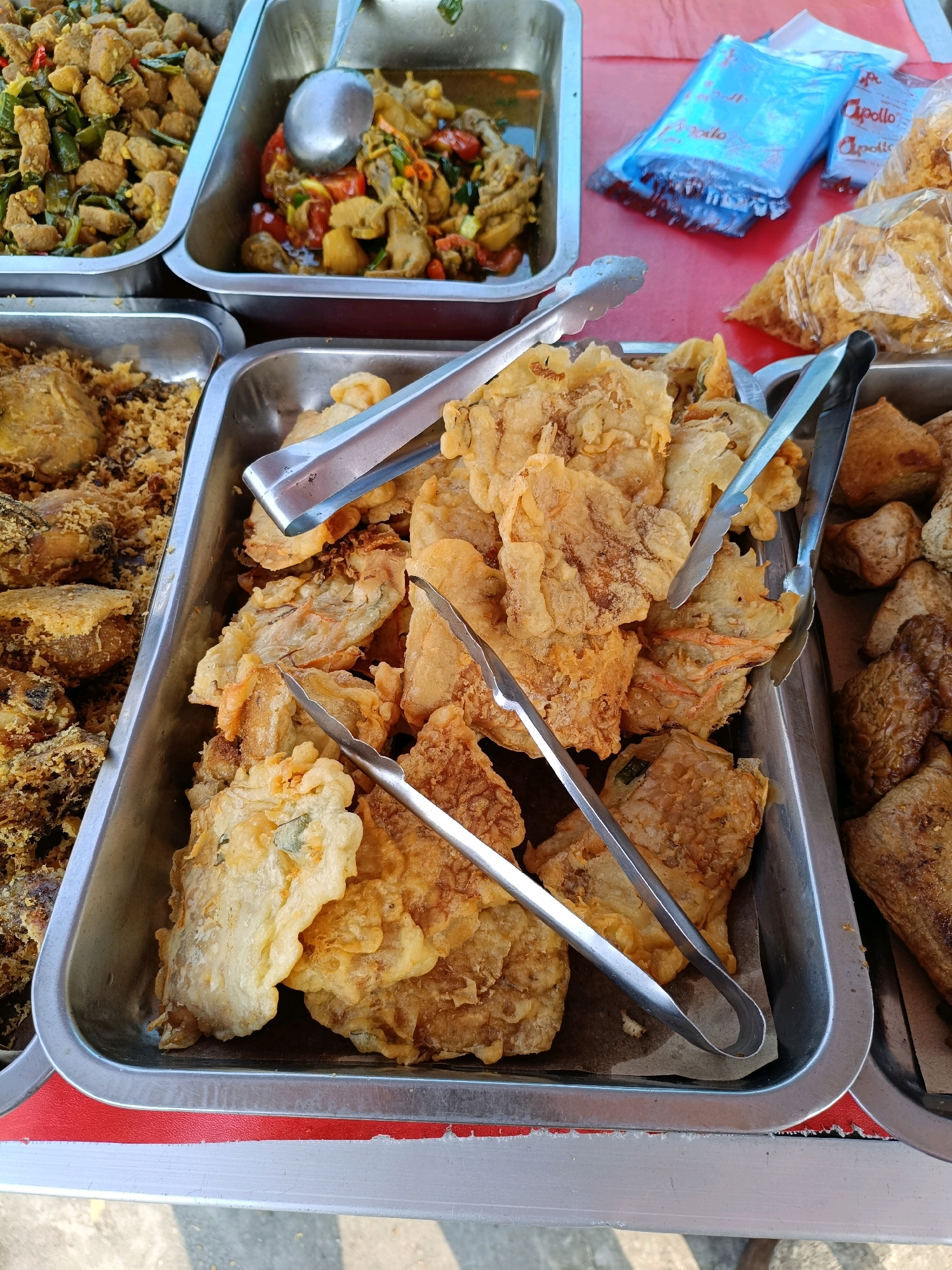 An overhead, close-up shot captures a street food stall in Maguwoharjo, Indonesia, featuring a variety of fried Indonesian dishes. The foreground is dominated by a rectangular metal tray filled with golden-brown fried tempeh and other battered items. Metal tongs are placed on top of the food, one set on the left and another on the right, indicating they are ready for serving. Behind this tray, a second metal tray holds more fried food, appearing drier and perhaps breaded. To the left, a third tray contains a dish with chunks of meat and vegetables in a sauce, with a serving spoon resting inside. Further back, a tray holds more fried food, and behind that, a clear plastic bag holds packaged snacks. The red tablecloth beneath the food and the pavement visible at the bottom edge suggest an outdoor setting. The lighting is natural and bright, casting subtle shadows. The image is focused on the food, with no people visible in the frame.