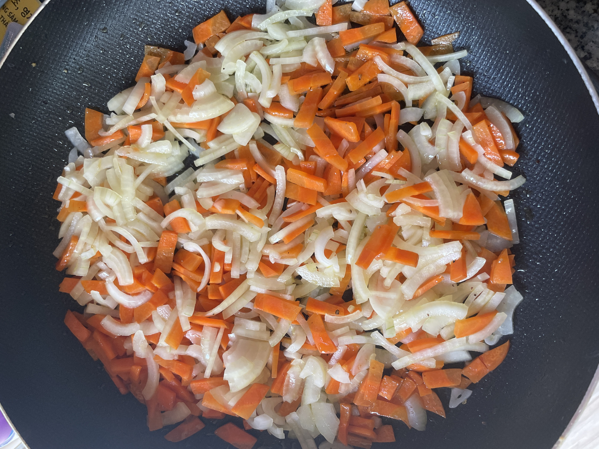 A close-up, overhead shot shows chopped carrots and onions sizzling in a black non-stick frying pan. The vegetables are cut into bite-sized pieces, with the carrots appearing as bright orange batons and the onions as translucent white slivers. The pan is coated with a thin layer of oil, and some of the vegetables have begun to soften and caramelize. The background is dark, highlighting the vibrant colors of the ingredients. There is no indication of the city of Phu Thuy Ward, Vietnam.