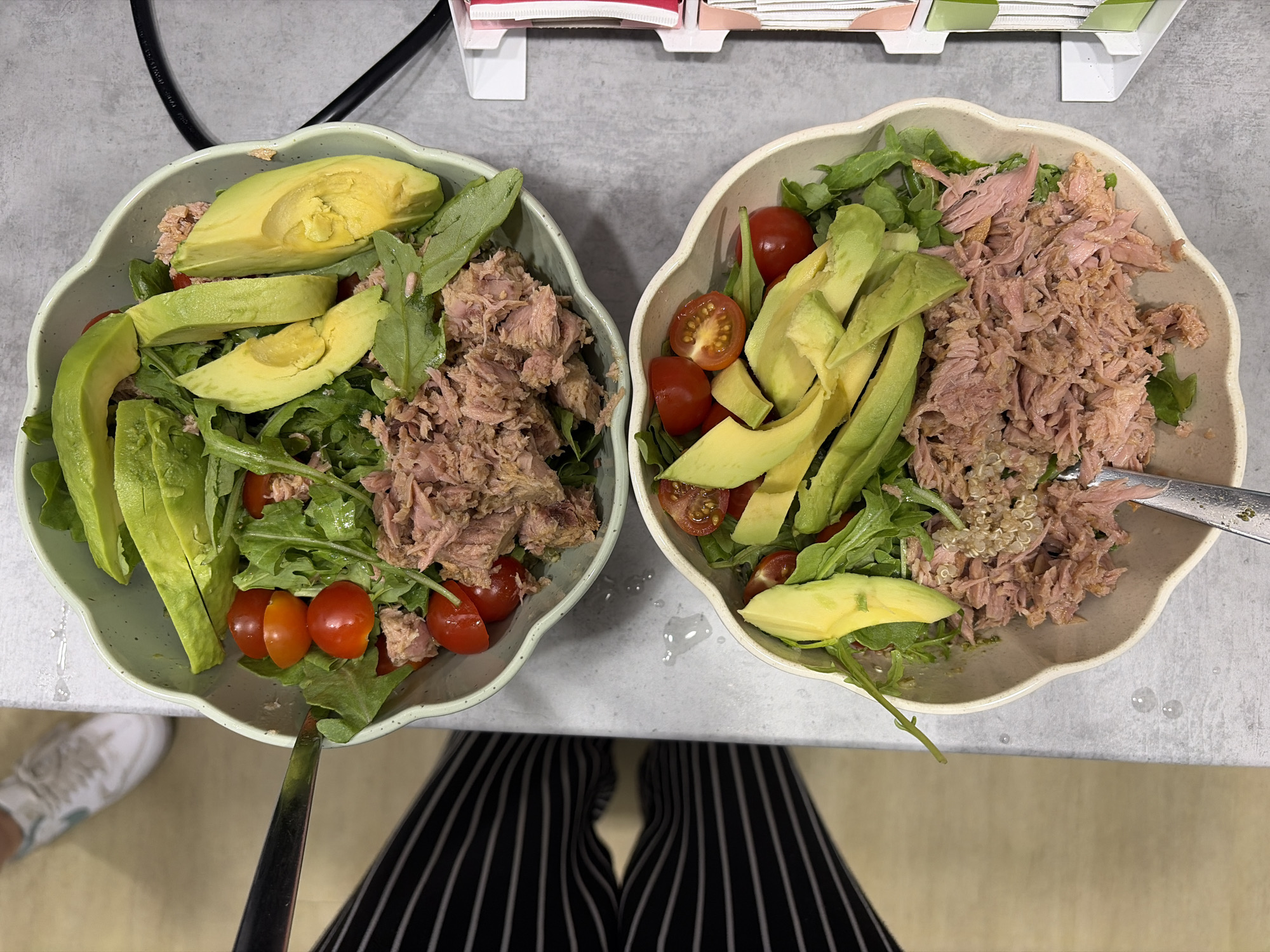 Two bowls of tuna salad with avocado and cherry tomatoes are presented on a gray surface. The salad in the top bowl contains arugula and shredded tuna, topped with sliced avocado and halved cherry tomatoes. A metal fork is partially visible in the top left corner of this bowl. The bottom bowl also contains shredded tuna, arugula, sliced avocado, and halved cherry tomatoes. Small white seeds, possibly quinoa, are mixed into the tuna in this bowl. A metal fork is visible in the bottom right corner of this bowl. In the background, a person wearing black and white striped pants and white sneakers can be seen. To the right of the bowls, a white dispenser with colorful compartments, possibly for utensils or napkins, is visible. The environment appears to be indoors, with water droplets on the gray surface between the bowls.
