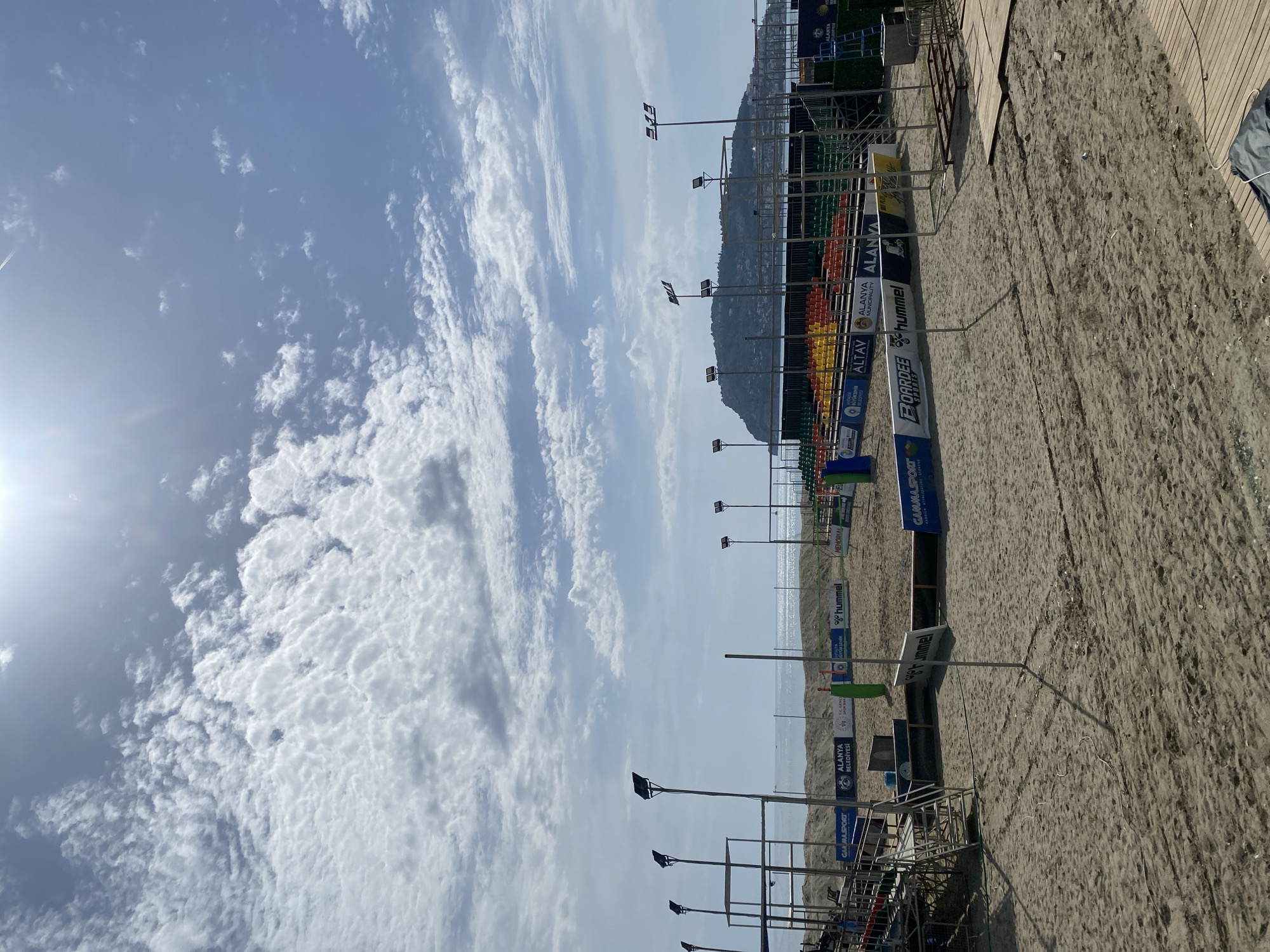 The image shows a beach volleyball venue in Alanya, Turkey. The foreground features a sandy beach with visible footprints. A spectator area with colorful seats is set up along the edge of the court. Advertising banners are erected along the sidelines, showcasing brands like "ALANYA Municipality" and "Hummel." Light posts are positioned throughout the area, presumably for nighttime events. In the background, a hazy horizon reveals the sea and a distant mountainous landscape. The sky is partly cloudy with the sun visible on the left side. A wooden walkway is seen on the right side of the venue.