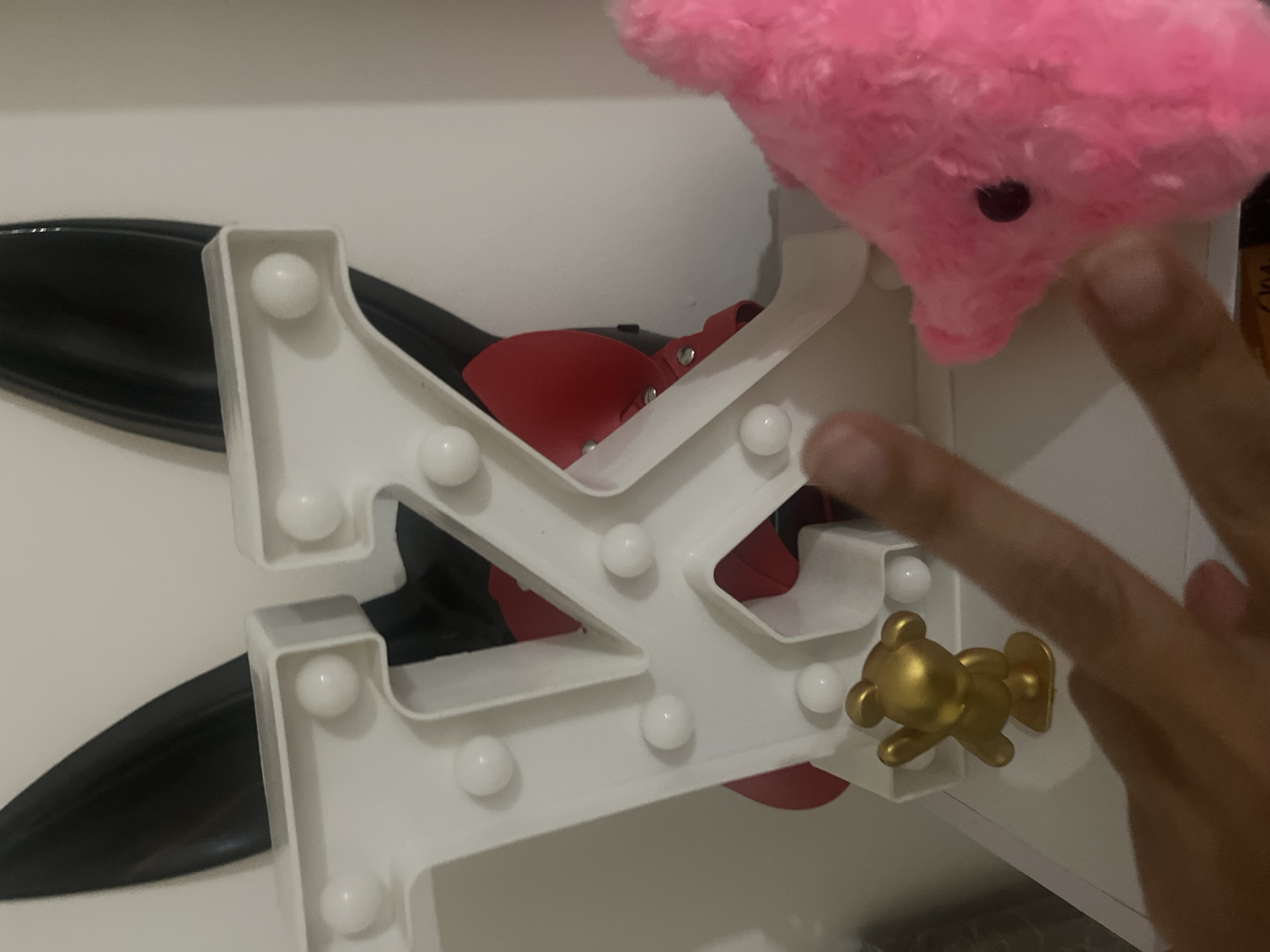 The image shows a close-up view of a white marquee letter "K" with lights. Behind the letter "K", there are black bunny ears. A small gold bear figurine is placed in front of the letter "K". To the right of the letter "K", a pink plush toy, possibly a pig, is visible. A person's hand is in the foreground, with fingers extended towards the letter "K". The background is a plain white wall. The scene appears to be indoors, on a shelf or flat surface. The image does not provide any specific location details that would allow for identification of Nha Trang Ward, Vietnam.