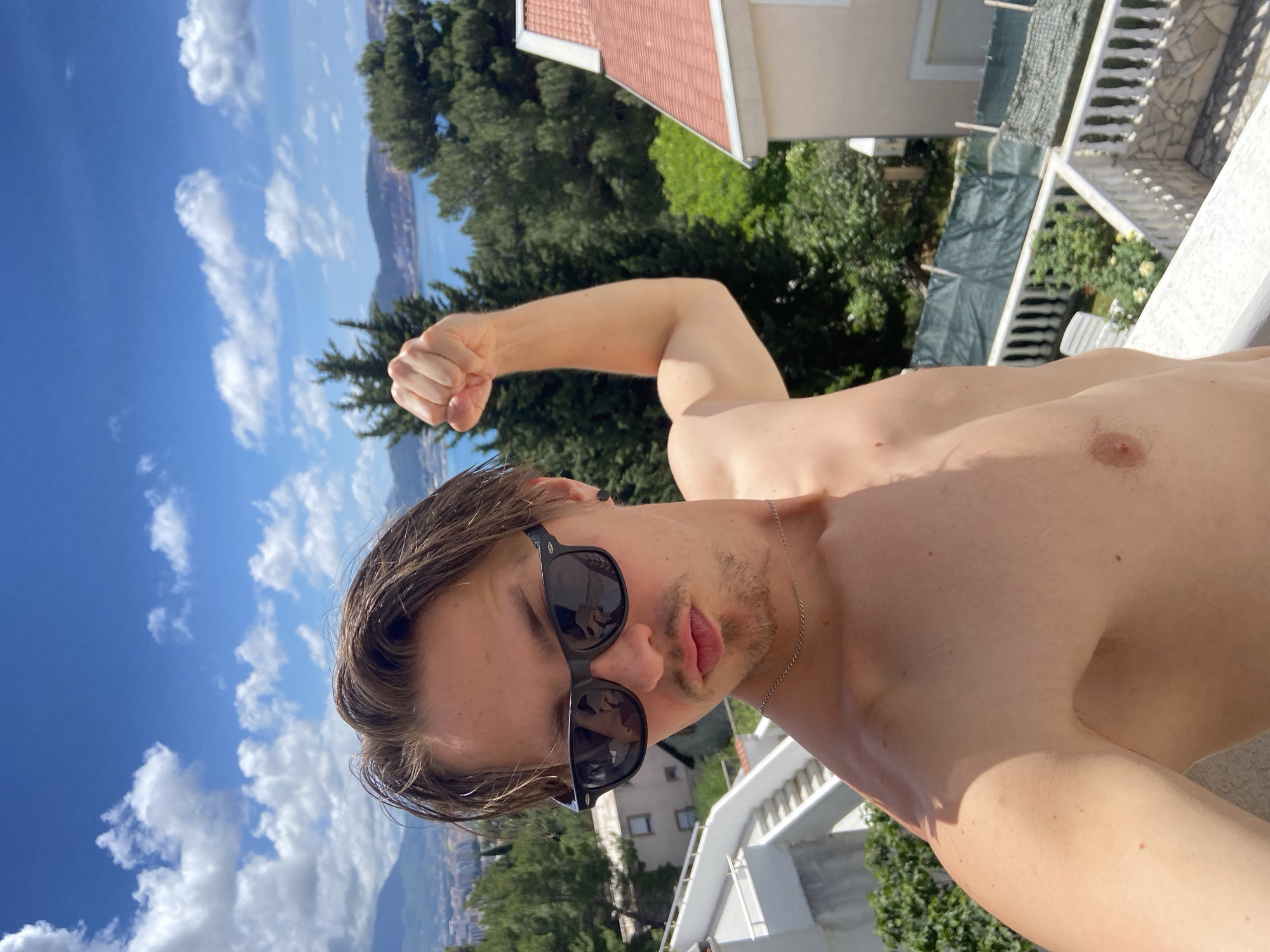 A shirtless man wearing sunglasses poses on a balcony in Šušanj, Montenegro. The man is fair-skinned with wet, medium-length brown hair, and light facial hair. He is flexing his bicep.
The background shows a coastal cityscape, green trees, and a blue sky with scattered clouds. A building with a red tile roof is partially visible. A white balcony railing and green vegetation surround the man.