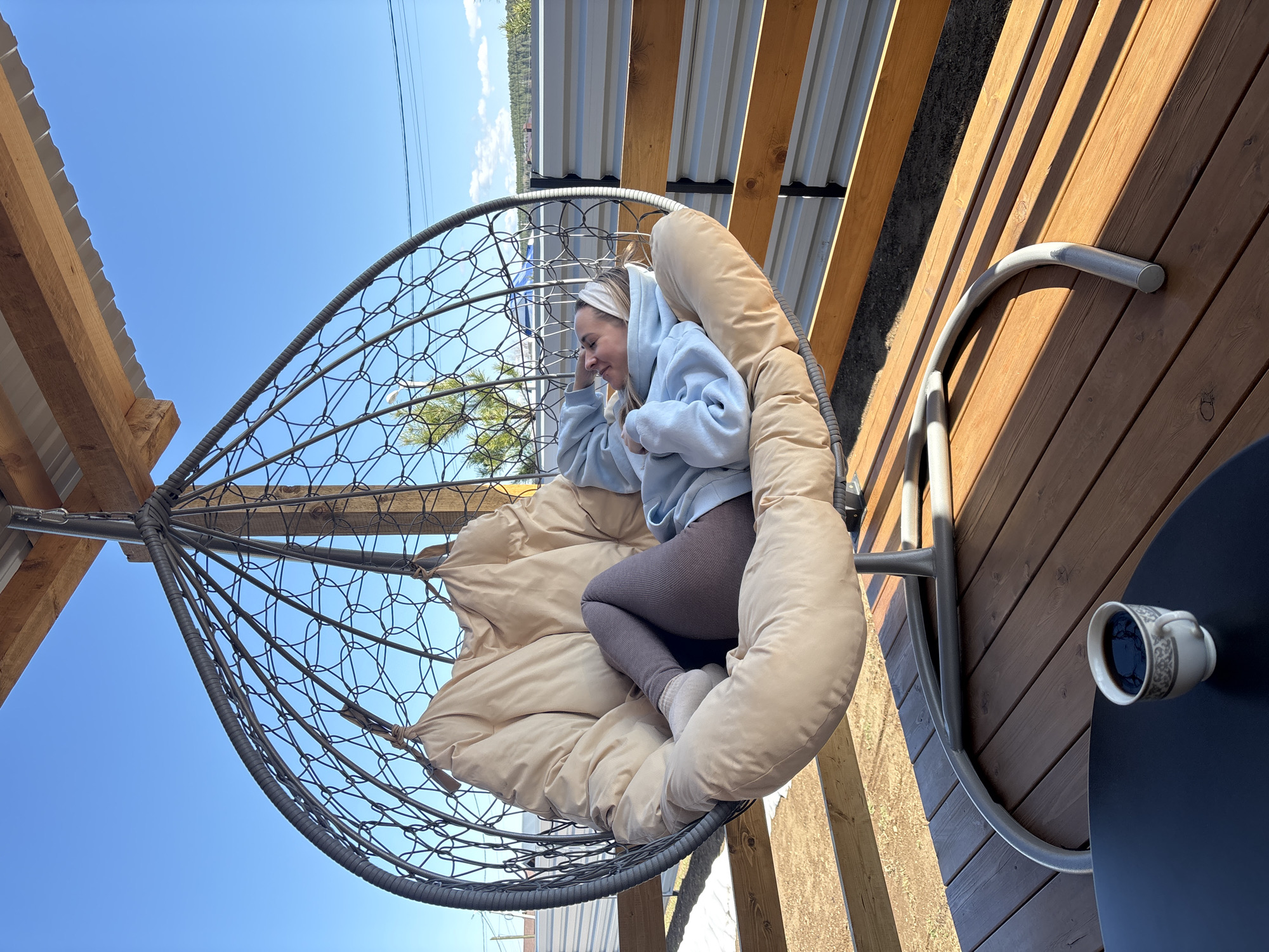 A young woman with blonde hair pulled back by a white headband is reclining in a hanging egg chair. She wears a light blue hoodie and brown leggings with white socks. The chair, lined with a thick beige cushion, sits on a wooden deck under a metal roof. A cup rests on a dark blue table beside the chair. In the background, through the chair's open netting, glimpses of trees and a bright blue sky are visible. The scene is bathed in sunlight. The location is identified as the city of Chaikovsky, Russia.
