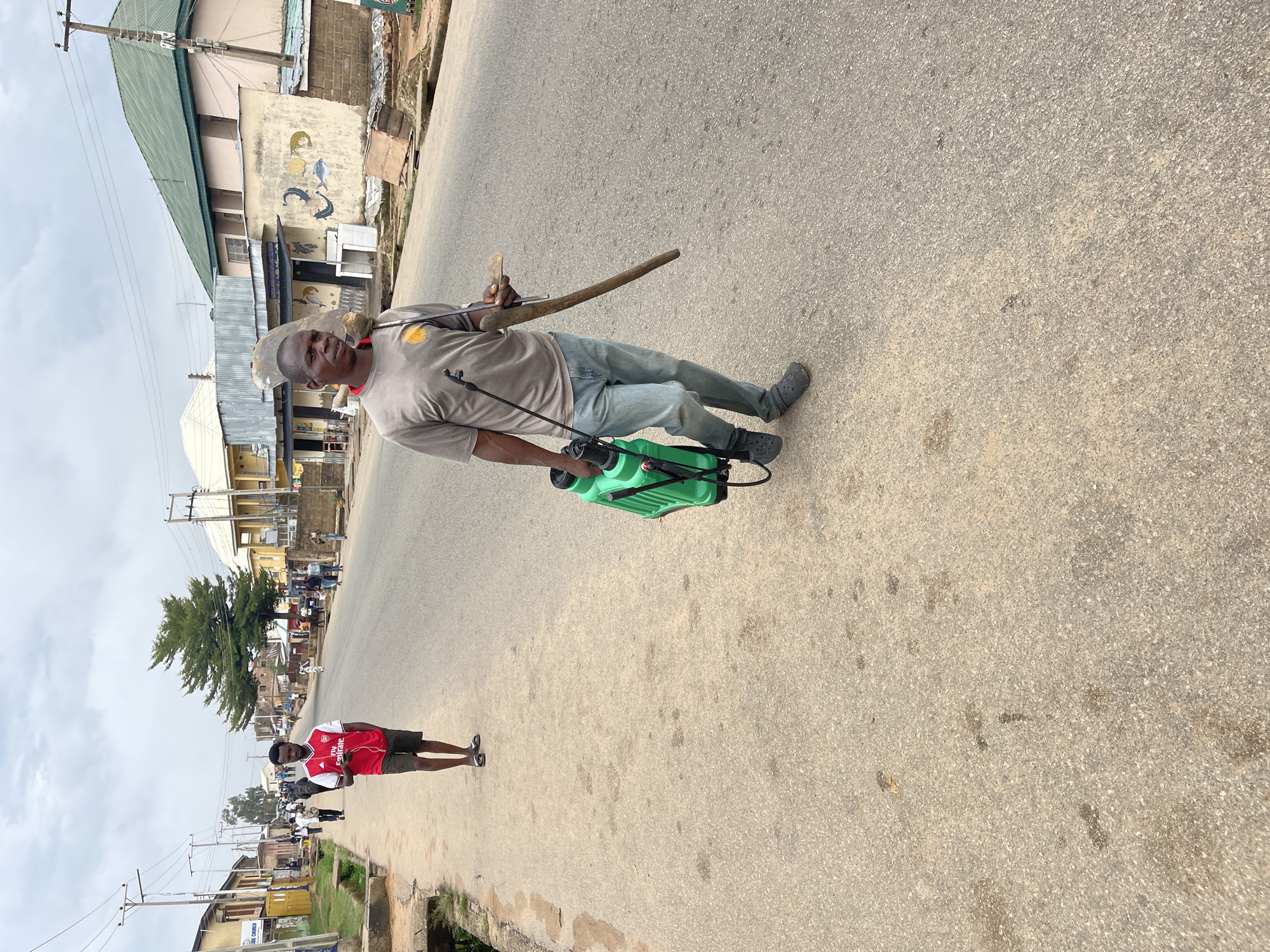The image shows two men on a paved road in Jos, Nigeria. One man wears a gray shirt, jeans, and black shoes, carrying a green sprayer and a wooden tool. He stands on the road, appearing to be spraying. The second man is further down the road, wearing an Arsenal soccer jersey, shorts, and sandals. He is standing near a sidewalk. Buildings and utility poles line the street in the background. The sky is overcast.