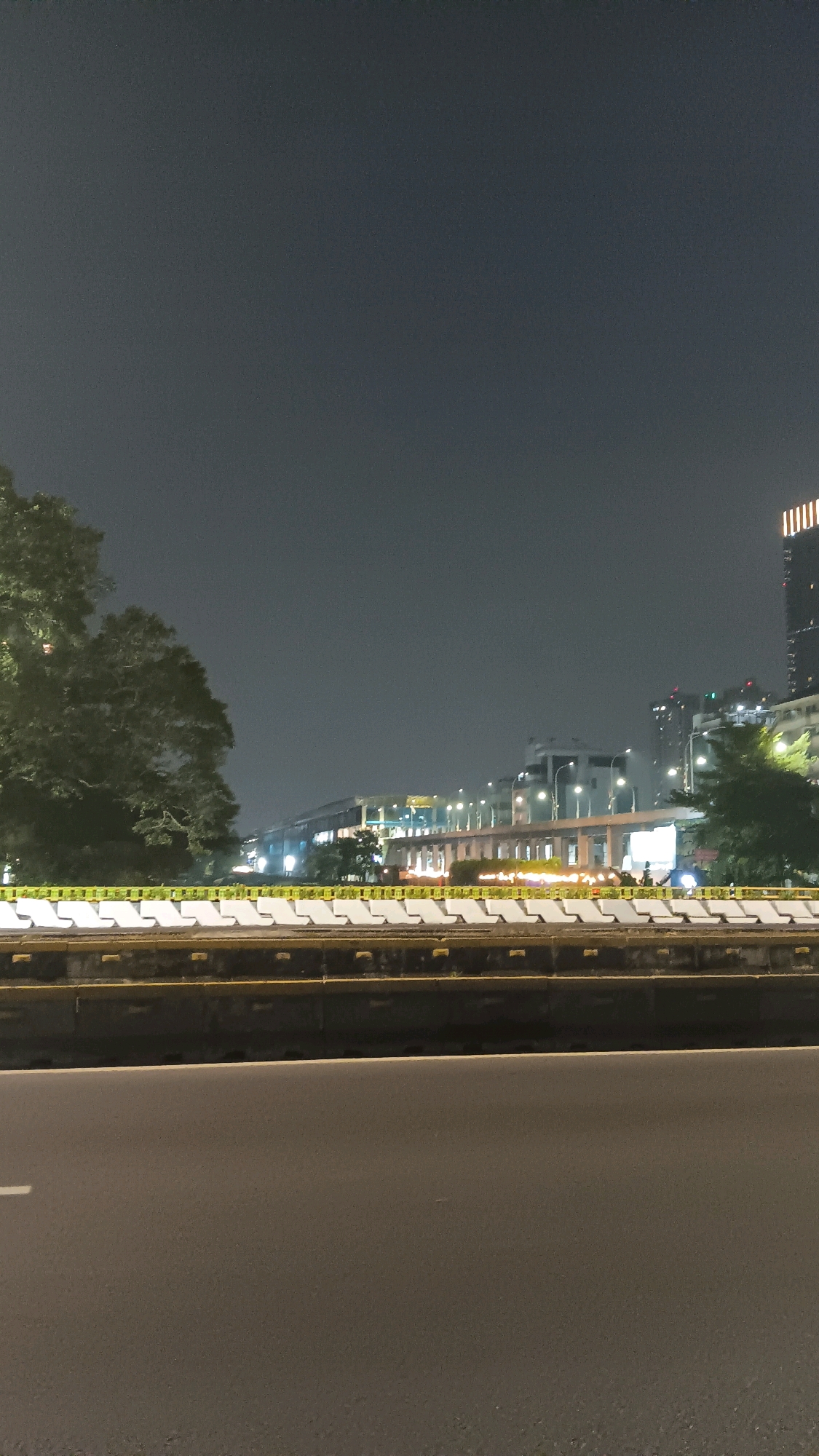 This is a nighttime photograph of an elevated roadway or structure in Central Jakarta, Indonesia. The foreground shows a dark asphalt road surface. Behind the road, there is a concrete barrier with white blocks and yellow detailing. Beyond the barrier, a long, illuminated structure with multiple levels of lights stretches into the distance. This structure appears to be an elevated train line or highway. In the background, city buildings and trees are visible against a dark, cloudy sky. The overall scene suggests an urban environment at night.