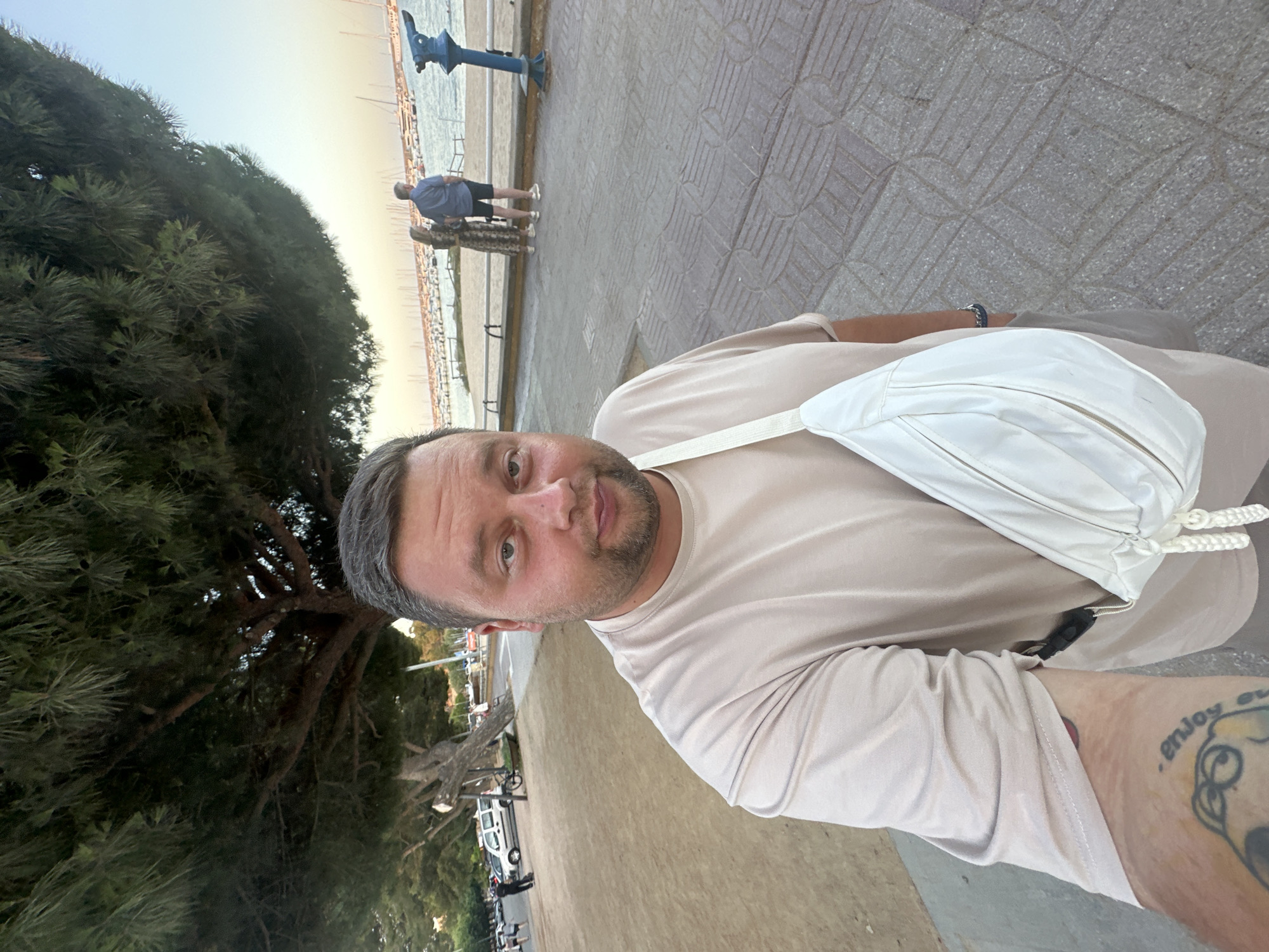 In Sant Feliu De Guíxols, Spain, a man is taking a selfie with a white fanny pack across his chest. He is wearing a beige t-shirt and has tattoos on his left arm. In the background, a tree with long, green needles overhangs the scene, with a paved walkway and a sandy area. Further in the background, two people stand near a large blue viewing scope overlooking a harbor filled with docked boats. The sky is clear and bright, indicating it is daytime.