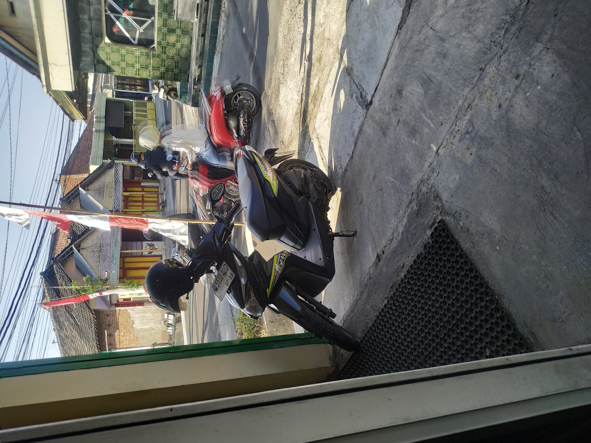 In Batan, Indonesia, a street scene unfolds under a clear sky, bathed in sunlight that casts sharp shadows on the ground. In the foreground, a black scooter is parked, its handlebars adorned with a black helmet. A black rubber mat lies in front of the scooter, partially on a concrete surface. Further down the street, a red scooter is visible with two individuals on it, both wearing helmets. The person in front has a white helmet and a white shirt, while the person behind wears a black helmet. Behind them, several houses with red tile roofs and painted facades suggest a residential area. Indonesian flags, red and white, are prominently displayed, one on a pole near the houses and another partially visible on the right side of the image. The presence of overhead wires indicates an urban or semi-urban setting. The overall impression is a snapshot of daily life in an Indonesian town.