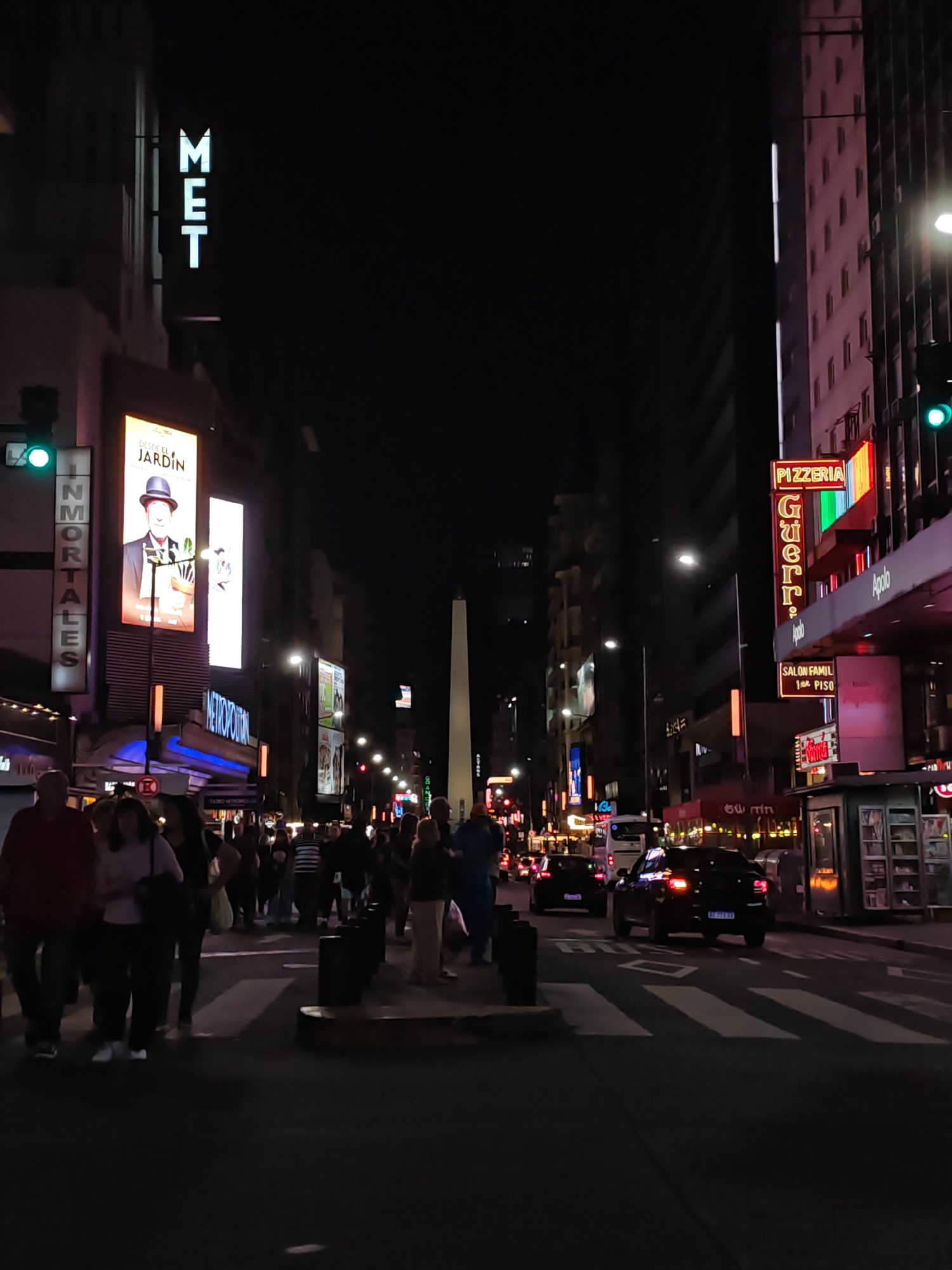 This is a nighttime street scene in Buenos Aires, Argentina. The iconic Obelisco monument is visible in the distance, centered within the street's perspective. A crowd of people is crossing the street at a zebra crossing. The street is illuminated by streetlights and neon signs. On the left side of the street, the vertical "MET" sign is visible atop a building, next to a large advertisement for "Desde El Jardin," and "Inmortales". On the right, there is a Pizzeria called "Guerrin" and an "Apolo" sign, along with smaller storefronts and a bus stop structure. Several vehicles are on the street, including at least two dark-colored cars. The overall impression is that of a lively, urban environment at night.